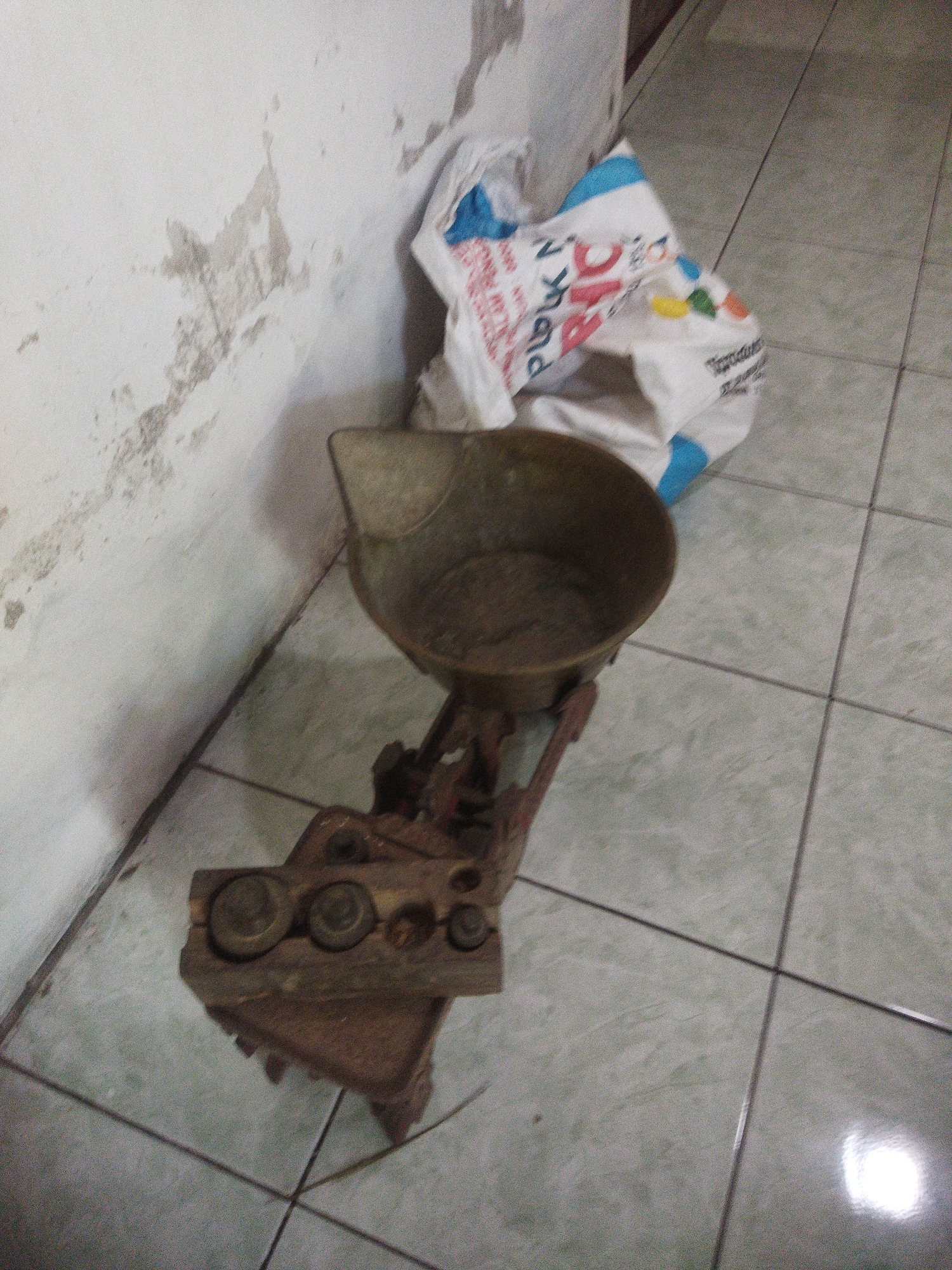 In a dimly lit room with tiled flooring and a distressed white wall, an old brass balance scale sits prominently. The scale features a round, shallow bowl at the top, which appears to be empty. Below the bowl, a red metal arm is connected to a base where several brass weights are arranged on a wooden tray. To the right of the scale, a crumpled white plastic bag with blue and red lettering is partially visible. The overall scene suggests a storage area or an unused corner of a room.
