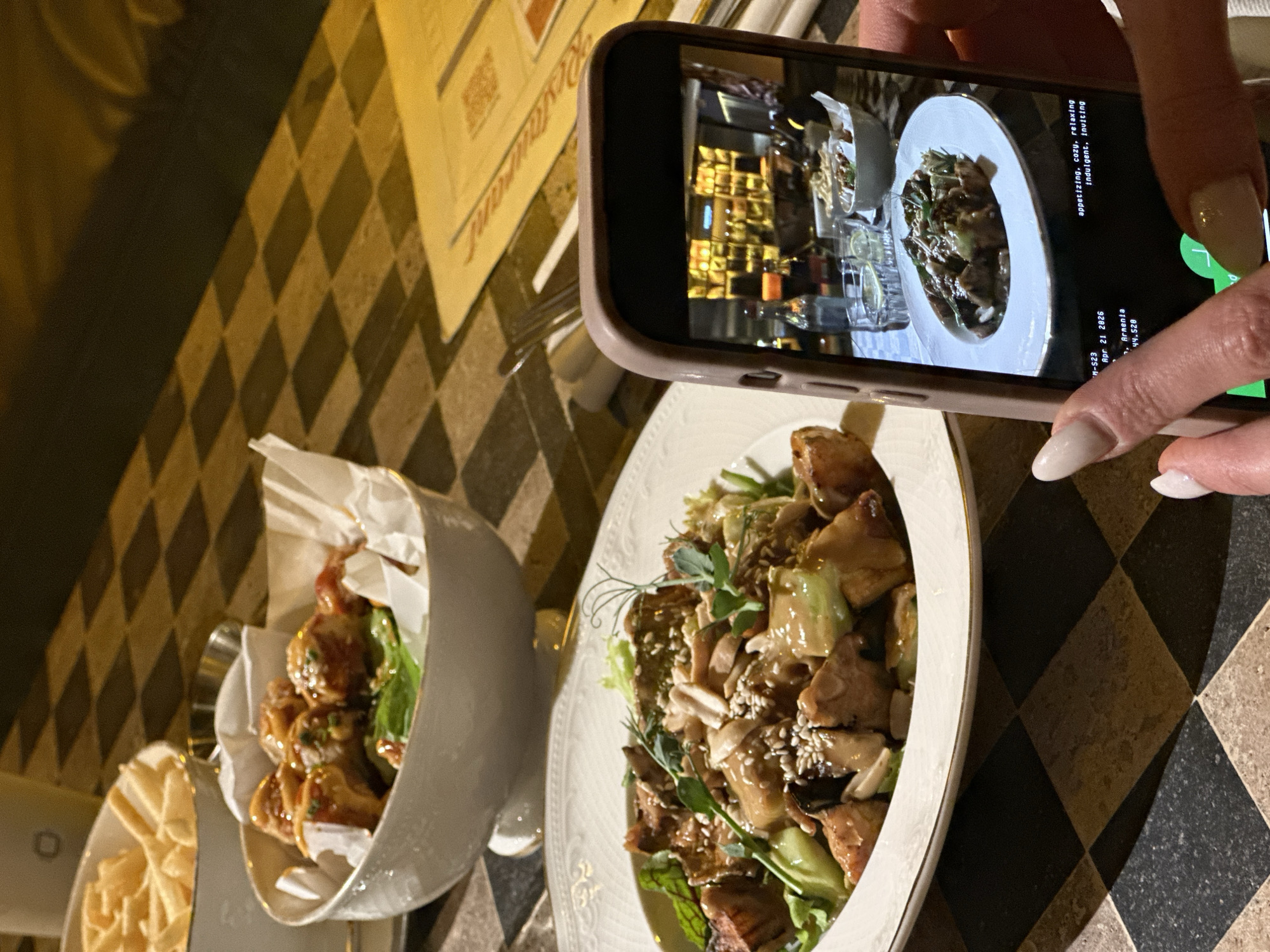 A person is capturing a photo of their meal with a smartphone in a dimly lit restaurant in Yerevan, Armenia. The right hand, with light-colored manicured fingernails, holds the phone vertically, aiming it at a plate of food. The phone screen displays the image being captured, or a previously taken photo, showing a close-up of a dish with reflections of the restaurant interior in the background.
On the table, there are two dishes. In the foreground, a white, ornate plate holds a main course composed of what appears to be pieces of chicken or meat, green vegetables (possibly bok choy or similar), topped with peanuts or almonds, sesame seeds, and fresh green sprouts. To the left, another dish is served in a stylish, possibly metal, container lined with paper, containing smaller fried items or crisps, next to a small bowl of what looks like thin crackers.
The table features a distinctive black and light-colored checkered pattern. A menu or placemat, with the word "Restaurant" visible, is partially under the phone.
Visible text on the phone screen includes: "M-S23", the date "Apr 21 2026", "Arnenta" (likely Armenia), and "44.520" (possibly coordinates). Descriptive adjectives also appear: "appetizing, cozy, relaxing, indulgent, inviting," setting a positive mood for the dining experience. The dim lighting suggests an evening meal, contributing to a cozy ambiance.