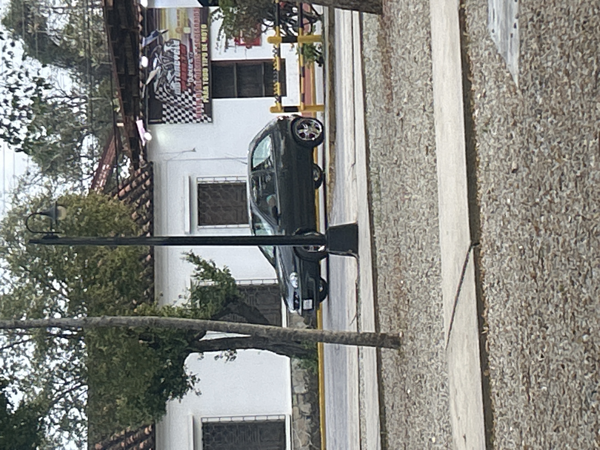 The image shows a street scene in Santa Ana, Venezuela. In the foreground, there is a sidewalk made of concrete slabs and gravel. A tree trunk runs vertically through the left side of the frame, partially obscuring a lamppost. In the middle ground, a dark-colored hatchback car is driving on the road. The car has custom chrome wheels. Behind the car, there is a yellow and black striped barrier and a white building with a red tile roof. A sign is visible on the building, featuring a checkered flag and the word "MOTORCYCLE" along with text in Spanish. The background consists of trees and foliage.