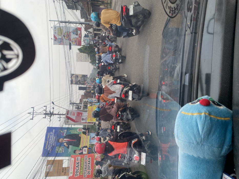 The image depicts a congested street scene in Mayong, Jepara Regency, Central Java, Indonesia, observed from the interior of a stationary vehicle during daylight under an overcast sky. Numerous individuals are present, primarily operating or riding as passengers on motorcycles, most wearing helmets of varying colors (red, blue, black, brown). The motorcycles are densely packed on the asphalt road, facing away from the viewer, with their taillights illuminated. Visible license plates include prefixes common to Central Java. The foreground shows the vehicle's dashboard, with a blue plush toy resting upon it. Overhead, a complex network of electrical wires and utility poles dominates the sky. Buildings line both sides of the street, with a series of large banners prominently displayed on the left. These banners include a large blue political advertisement for the PKB (National Awakening Party) congratulating its 27th anniversary and a red protest banner stating "TOLAK !!! PERUSAHAAN PETERNAKAN BABI DI JEPARA" (REJECT !!! PIG FARMING COMPANY IN JEPARA). Other commercial advertisements are visible further down the street.