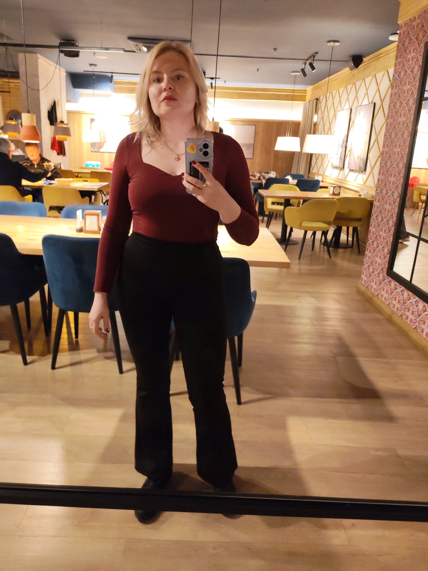 A full-length mirror selfie shows a woman with blonde hair, wearing a long-sleeved burgundy top and black pants. She is holding a smartphone with a case in her right hand, and her left hand is at her side. The background reveals a restaurant interior with wooden floors, tables, and chairs. One section has blue chairs, while others have yellow. There is patterned wallpaper and artwork on the walls. Other people are visible seated at tables in the background. There is no information provided to confirm the location.