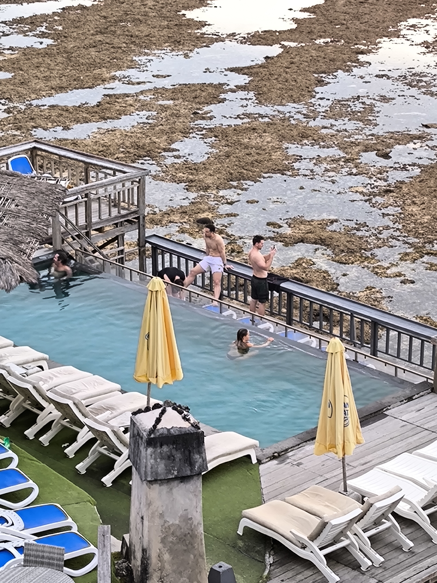 The image depicts an infinity pool scene located in Uluwatu, Indonesia. The pool is surrounded by lounge chairs with yellow umbrellas, some on artificial turf, and others on a wooden deck. Several people are in or near the pool. One person is swimming, and others are standing near the edge. Two men appear to be taking photos. Beyond the pool's edge, a large expanse of rock formations and tidal pools are visible.