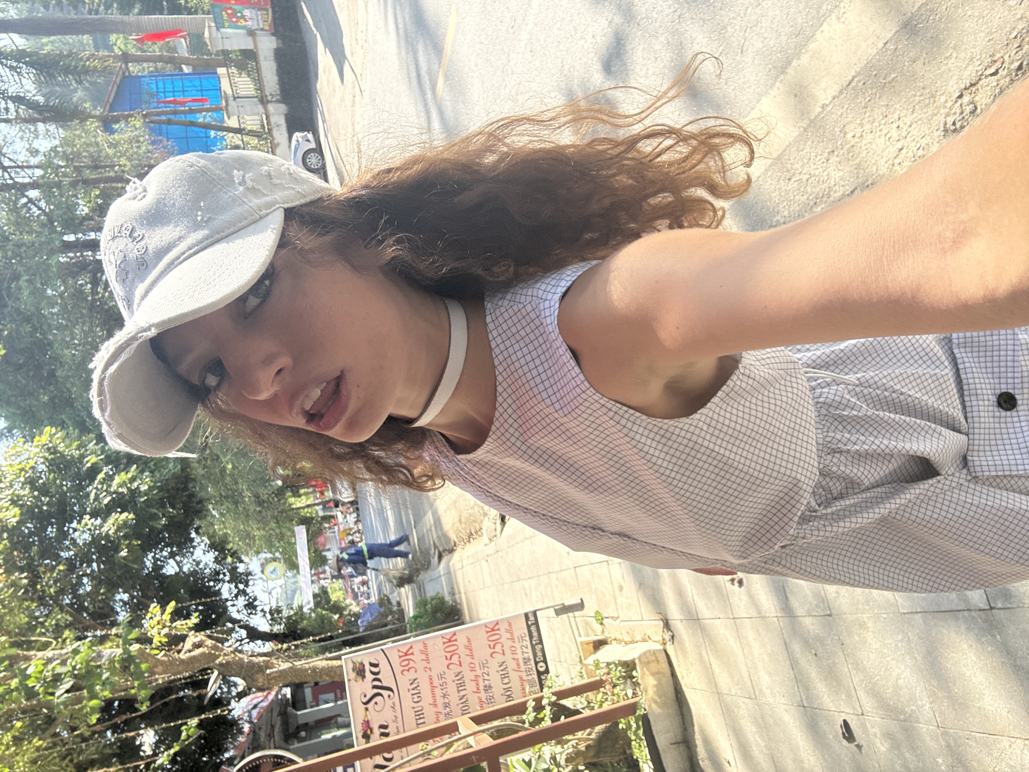A woman is taking a selfie outdoors on a sunny day. She wears a distressed grey baseball cap and a white-and-grey checkered dress with a white choker necklace. Her curly brown hair is partially blowing in the wind. The setting appears to be a street in Bắc Hà, Vietnam, as indicated by the spa sign in the background with prices in Vietnamese currency. The sign advertises spa services with prices in both Vietnamese Dong and US dollars, suggesting a tourist area. The street is paved and there are trees and other buildings visible in the background. People are walking in the distance.
