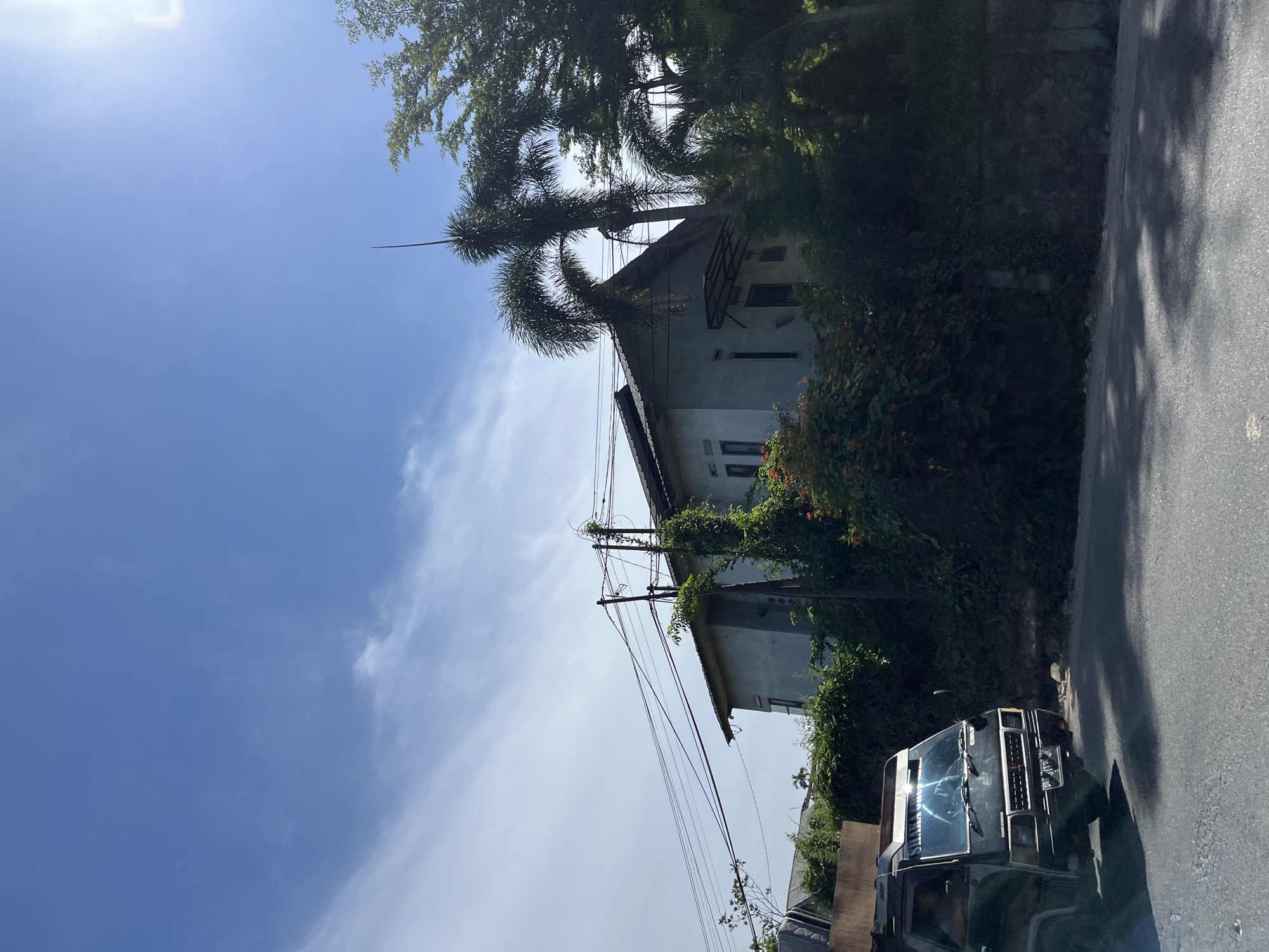 This is a vertical, outdoor, daytime photograph taken in Bukitrata, Indonesia. The scene depicts a street with a dark-colored utility vehicle parked on the right side, facing away from the viewer. The vehicle has a license plate with some visible text, though it is partially obscured. To the left of the vehicle, there is a densely vegetated area with trees and flowering plants, some of which are encroaching on a light-colored building. The building appears to be a house with windows and a sloped roof, and it is partially covered in vines. Several utility poles with numerous wires run along the side of the street and past the building. The sky is clear blue with some wispy white clouds, indicating good weather. Sunlight creates dappled shadows on the asphalt road, suggesting it is a bright, sunny day. The overall impression is a tropical or subtropical residential setting.