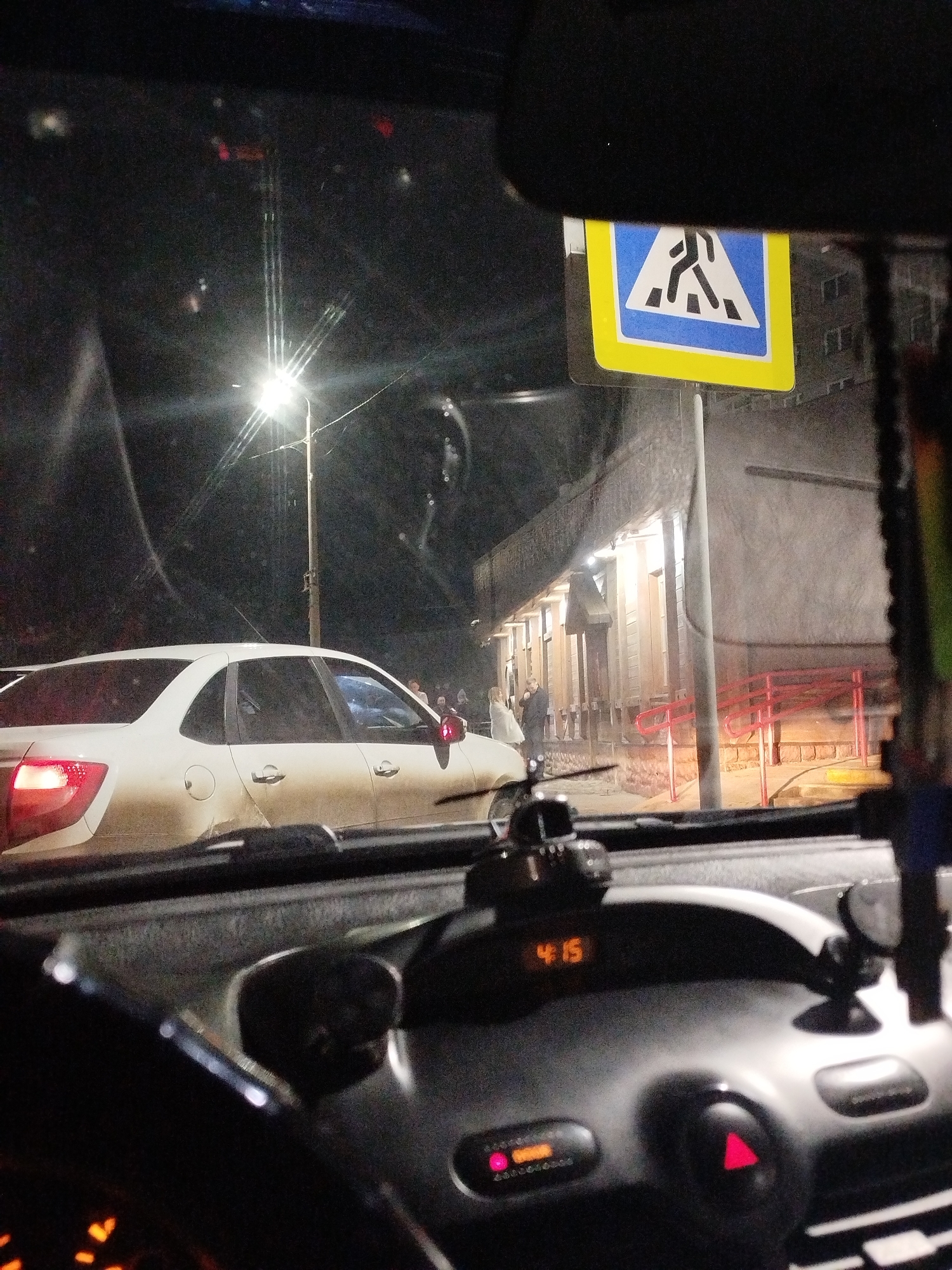 The image depicts an urban street scene at night, viewed from the interior of a stationary vehicle. The vehicle's dashboard features a digital clock displaying "4:15" and an illuminated red hazard light button. A light-colored sedan, possibly a Lada Granta, is visible in the midground, positioned on the street with its rear facing the viewer. Its left taillight is active, and the vehicle appears covered in dirt.
To the right of the sedan, a group of approximately four individuals stands on a sidewalk in front of a multi-story building. Two individuals are prominent, one in a light jacket facing another in a dark jacket. The building's ground floor features illuminated entrances and a red handrail leading to steps. A street light pole with a bright luminaire stands between the sedan and the building. A standard Russian pedestrian crossing sign, characterized by a blue square containing a white triangle with a black pedestrian symbol and a yellow border, is visible in the upper right portion of the frame. This scene is located in the city of Chaikovsky, Russia.