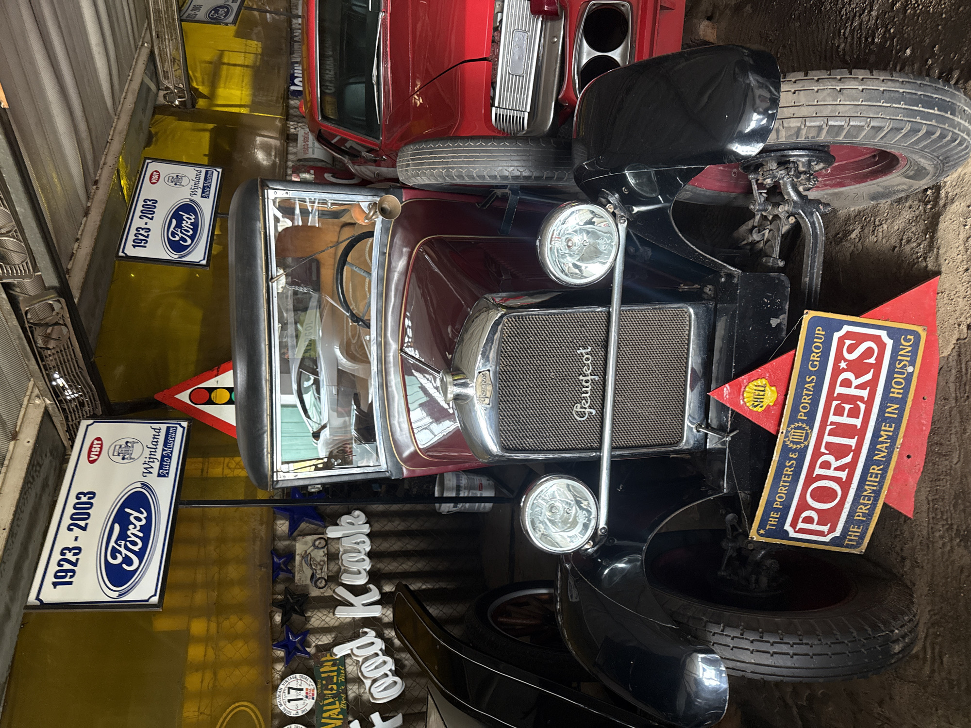 The image depicts a display of vintage cars and related memorabilia, likely within a museum or collection setting. Dominating the foreground is a classic Peugeot automobile, presented alongside a triangular sign for "The Porters & Portas Group" with "PORTER'S" prominently displayed, advertising it as "THE PREMIER NAME IN HOUSING". A Shell logo is visible on the same sign. Adjacent to the Peugeot are signs advertising "Ford" from "1923-2003" and “Wijnland Auto Museum”. Additional signage and decorative items, like a "Food Kiosk" sign and Valvoline marketing material, are visible in the background. Another vintage red car is partially visible above the Peugeot. The location is indicated on the Ford sign as "Wijnland Auto Museum", which can be found in Kraaifontein, South Africa. The scene appears to be inside a building with a metal ceiling.