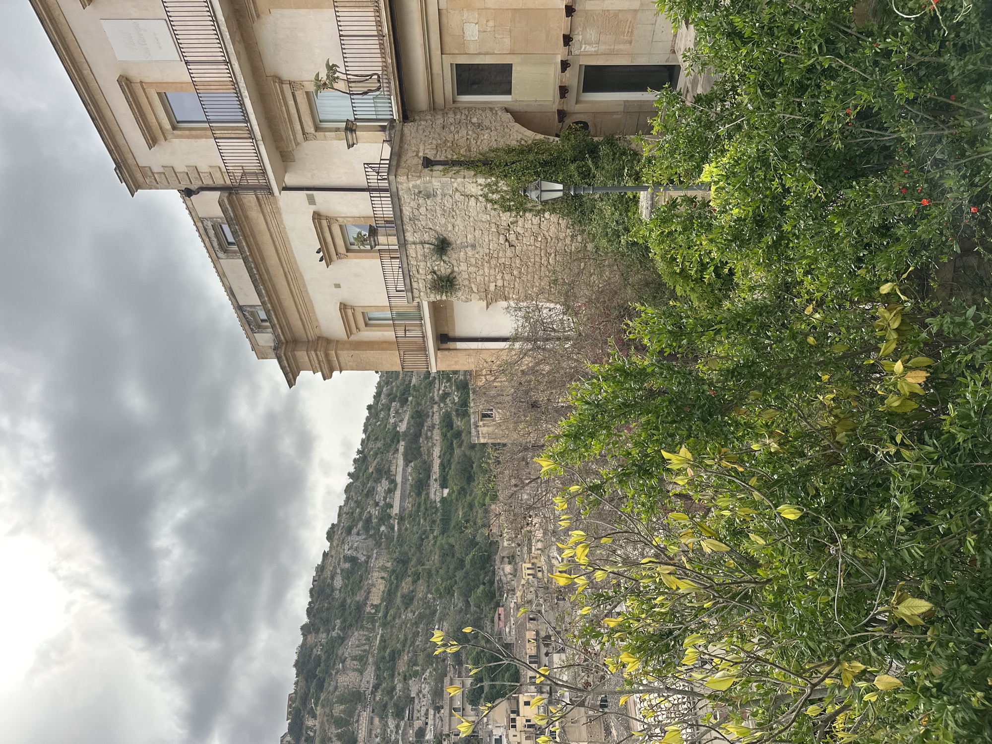 The image shows a view in Modica, Italy. A multi-story building with balconies and a stone facade stands on the left. A street lamp is attached to the stone facade. A green tree occupies the right side of the image. In the background is a mountain covered in vegetation and a cluster of buildings. The sky is cloudy and overcast.
