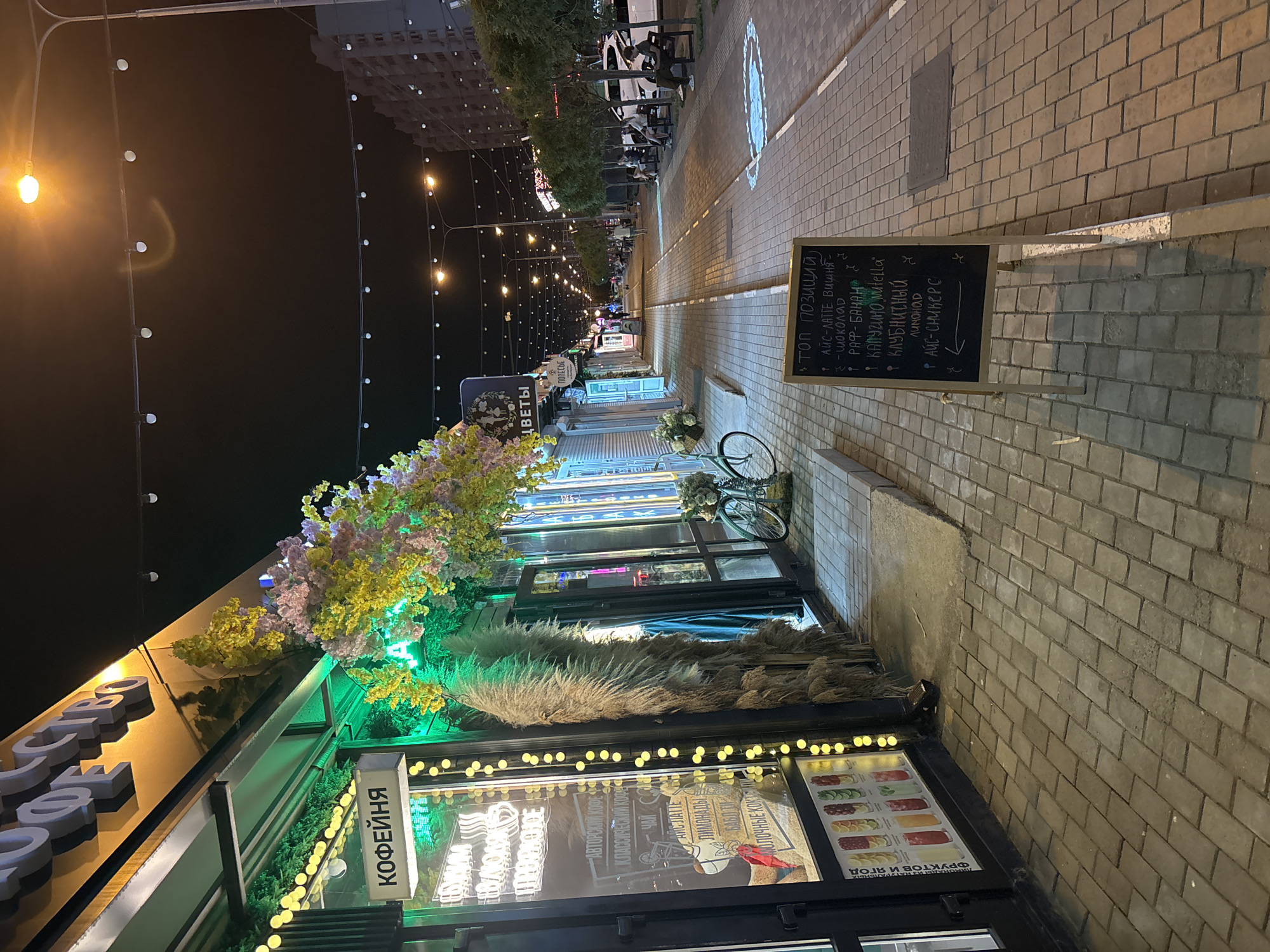 The image shows a street scene at night in Krasnodar, Russia. A coffee shop called "КОФЕЙНЯ" is prominently featured on the left side of the image. The shop has illuminated signs and decorations, including string lights and artificial floral arrangements. A bicycle is parked in front of the shop. A blackboard sign displaying a menu is placed on the sidewalk in front of the shop, listing items such as "АЙС-ЛАТТЕ ВИШНЯ," "ШОКОЛАД," "РАФ-БАНАН," "КАПУЧИНО Nutella," "КЛУБНИЧНЫЙ ЛИМОНАД," and "АЙС-СМЙКЕРС." The street is paved with brick pavers and lined with trees and benches. Several people are visible sitting on benches and walking along the street. Buildings are visible in the background.