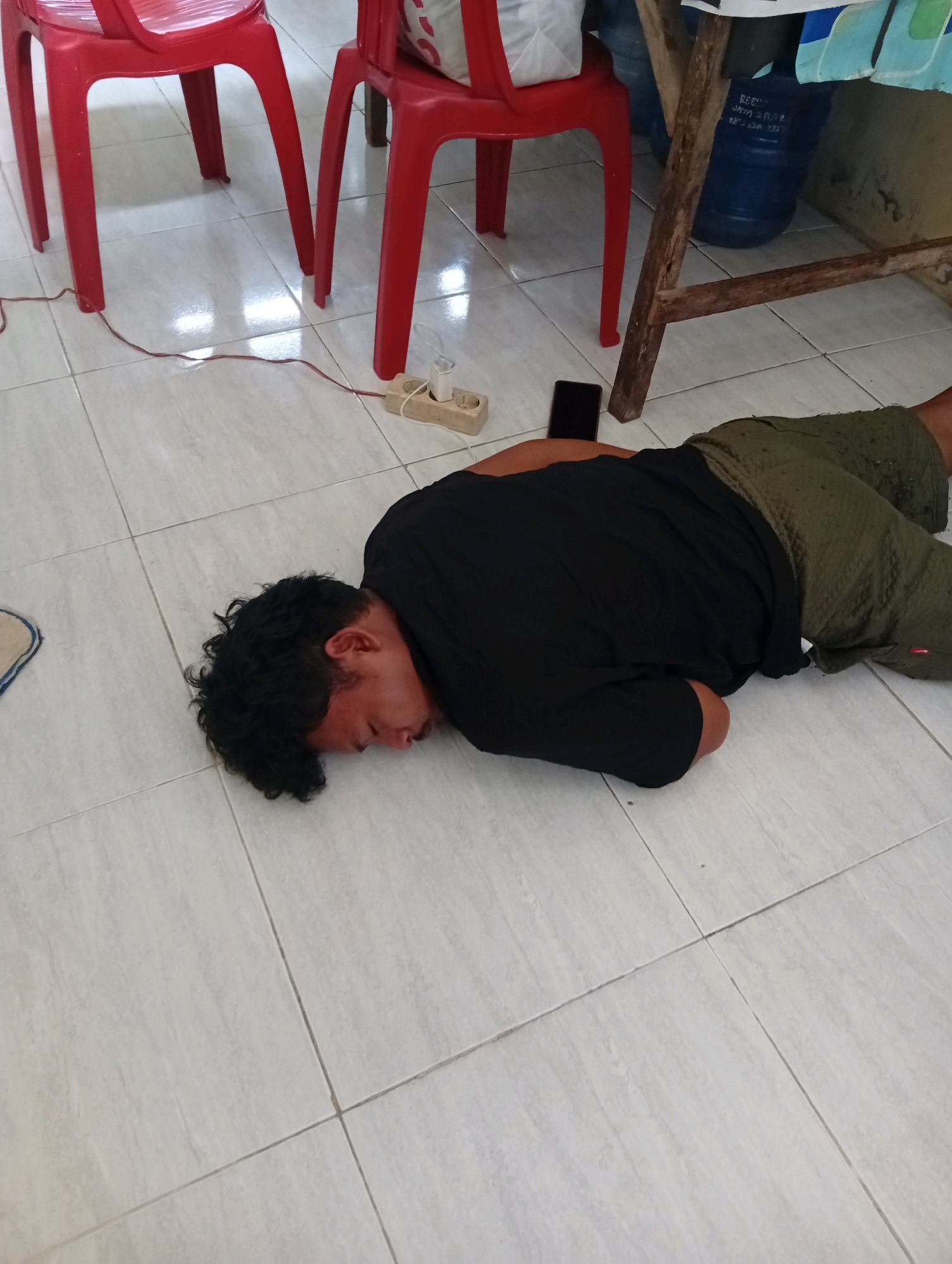 A young male with curly black hair is lying face down on a tiled floor. He is wearing a black t-shirt and olive green shorts. His right arm is bent, and his face is resting on the floor. A black smartphone is placed on the floor near his head. In the background, two red chairs are visible, and a wooden table leg is on the right side of the image. A power strip with a white plug is on the floor near the chairs. The scene appears to be indoors, possibly in Kualu, Indonesia, given the tiling and general appearance.