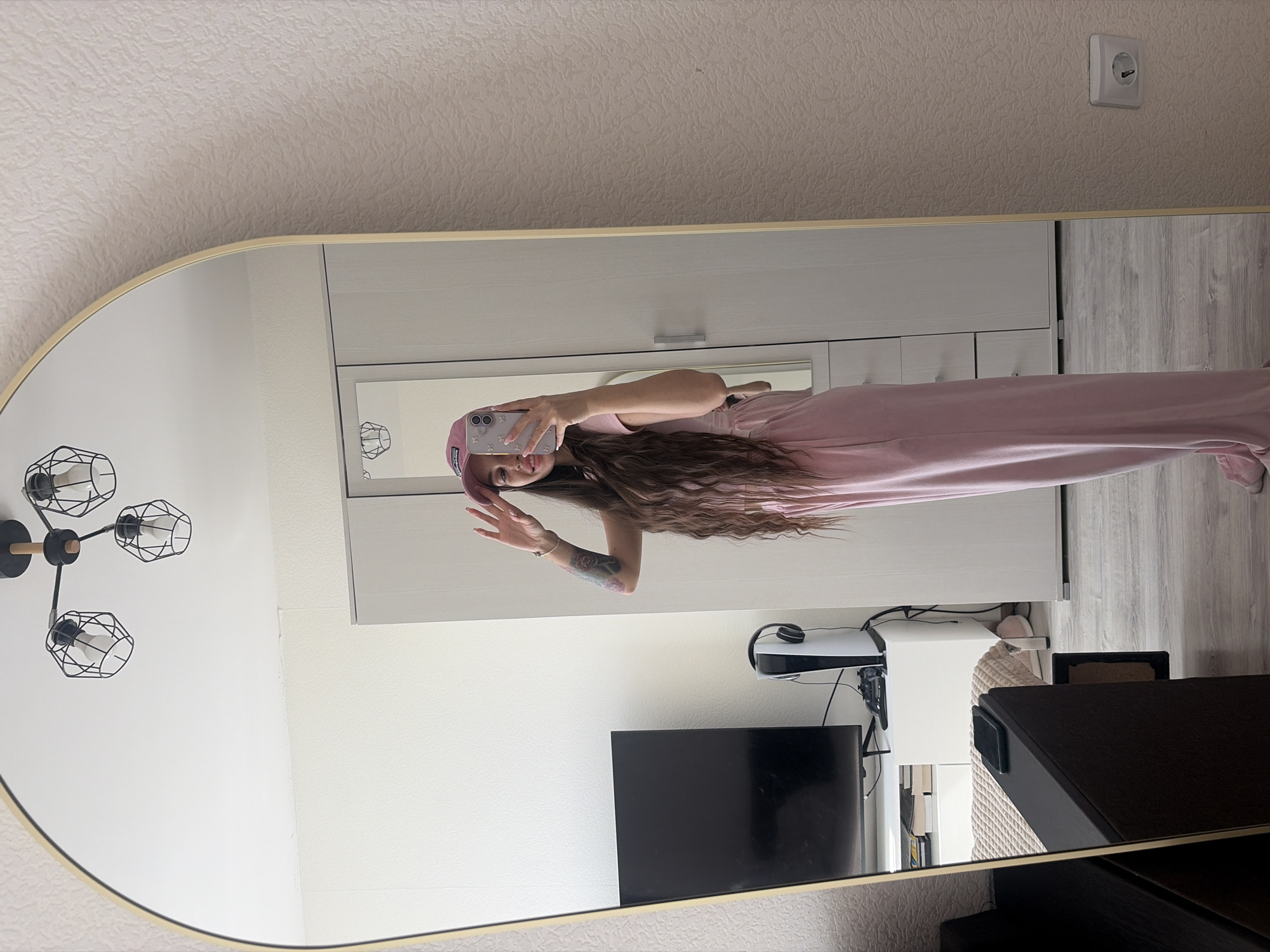 A woman in a pink outfit and matching cap is taking a selfie in a mirror. She has long, wavy brown hair and a tattoo on her left arm. In the background, there is a white wardrobe, a television, a PlayStation 5, and a black chair. The room appears to be located in Saint Petersburg, Russia based on the European style outlet.
