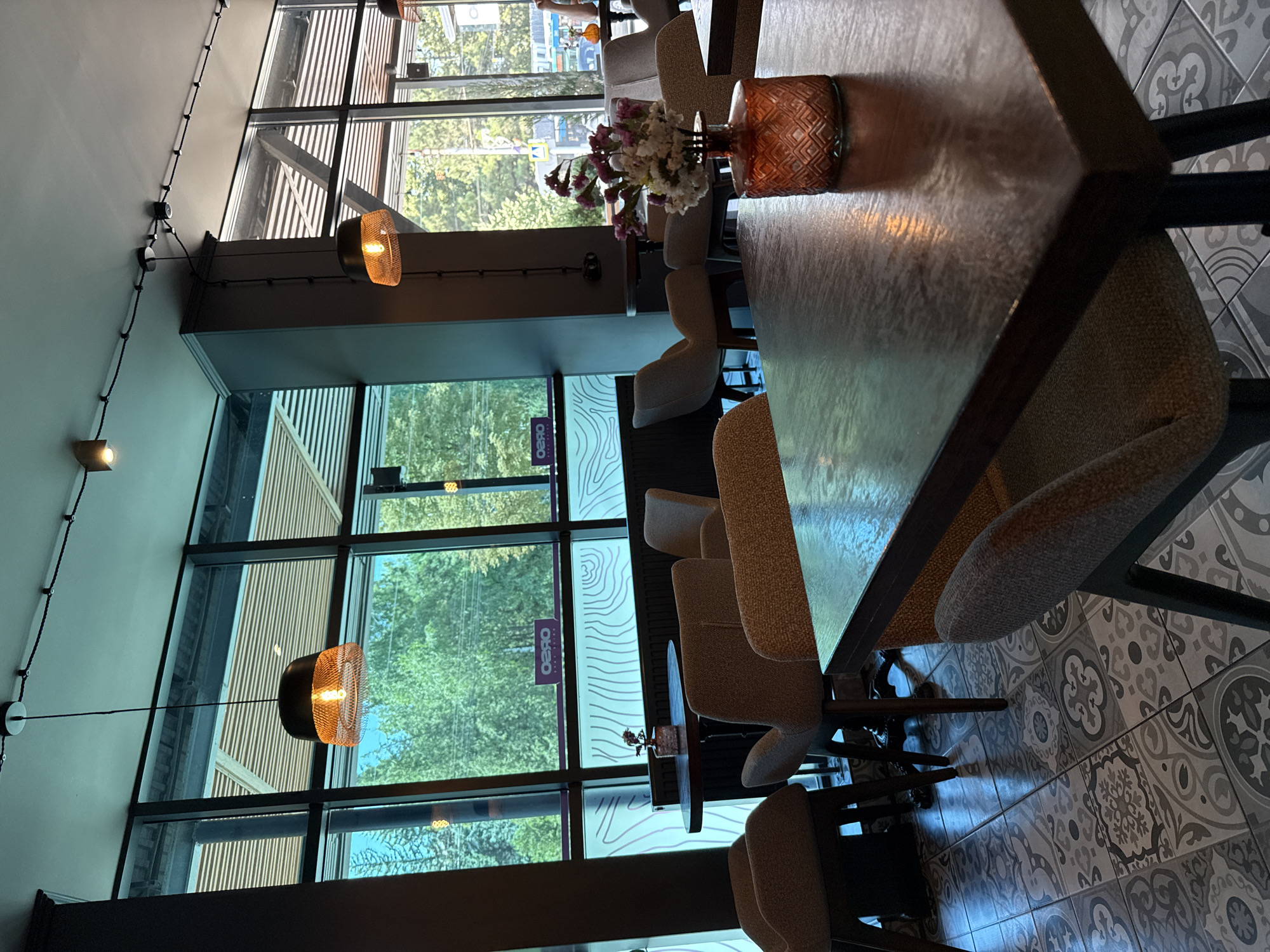 The image depicts the interior of a cafe in Bălți, Moldova, characterized by a modern and cozy ambiance. Large windows dominate the left and right sides of the frame, offering a view of lush green trees and a glimpse of the city street. The cafe is furnished with a variety of comfortable seating options, including upholstered chairs and armchairs in neutral tones. Several tables are strategically placed throughout the space, some with a polished wooden finish and others with a darker, more industrial aesthetic.
Decorative elements add to the charm of the cafe. A small vase filled with delicate purple and white flowers sits on one of the tables, its orange patterned glass base catching the light. Overhead, industrial-style pendant lights with mesh shades cast a warm glow. The floor is covered in an intricate patterned tile mosaic, adding a touch of visual interest.
In the background, through the windows, we can see streetlights and hints of traffic, suggesting the cafe is situated in an urban environment. A sign with "OBRO" in reversed lettering is visible on the window, likely indicating the name of the establishment. The overall impression is one of a well-designed and inviting space, suitable for relaxation or socializing. There are no people visible in the image.