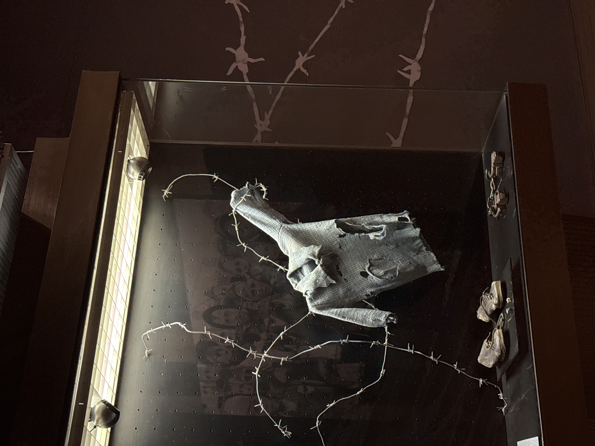 The image depicts an exhibit within a museum, likely a memorial. In the center, a tattered, light blue sweater hangs on a barbed wire structure. Below the sweater, a pair of worn leather shoes rests on a dark surface. To the right of the shoes, a set of metal handcuffs is displayed. The background of the exhibit features a blurred, monochrome photograph of a crowd of people, suggesting a historical context. The overall environment is dimly lit, with spotlights illuminating the artifacts. The exhibit is enclosed within a glass display case. The city of Kyiv, Ukraine is not visible in this image.