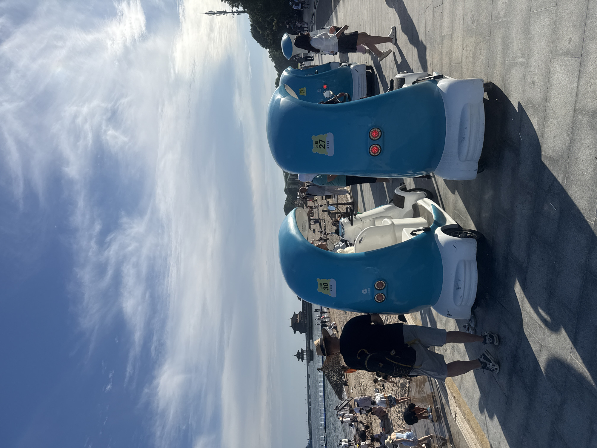 This is a photograph taken at a beachside promenade in Qinhuangdao, China, likely at Beidaihe's Zhonghai Beach. In the foreground, two blue and white electric recreational vehicles, designed to resemble a stylized wave, are parked on a paved walkway. One vehicle displays a "30" and Chinese characters on a yellow sticker, indicating it's a rental unit. The other has a "27" on a similar sticker.
A man wearing a straw hat, a dark t-shirt, and grey shorts stands facing away from the camera, with a backpack slung over his shoulder. His shadow is cast on the walkway. Further back, a crowd of people are visible on the sandy beach. Some are wading in the shallow water, while others are sitting or playing.
In the background, the sea stretches out to the horizon under a partly cloudy blue sky. Across the water, traditional Chinese-style pavilions are visible on a pier or connected to the shore by a bridge. The promenade is lined with trees on the right side of the image, and a tall telecommunications tower is also visible in the distance. The overall scene depicts a sunny day at a popular tourist destination, with people enjoying recreational activities by the sea.