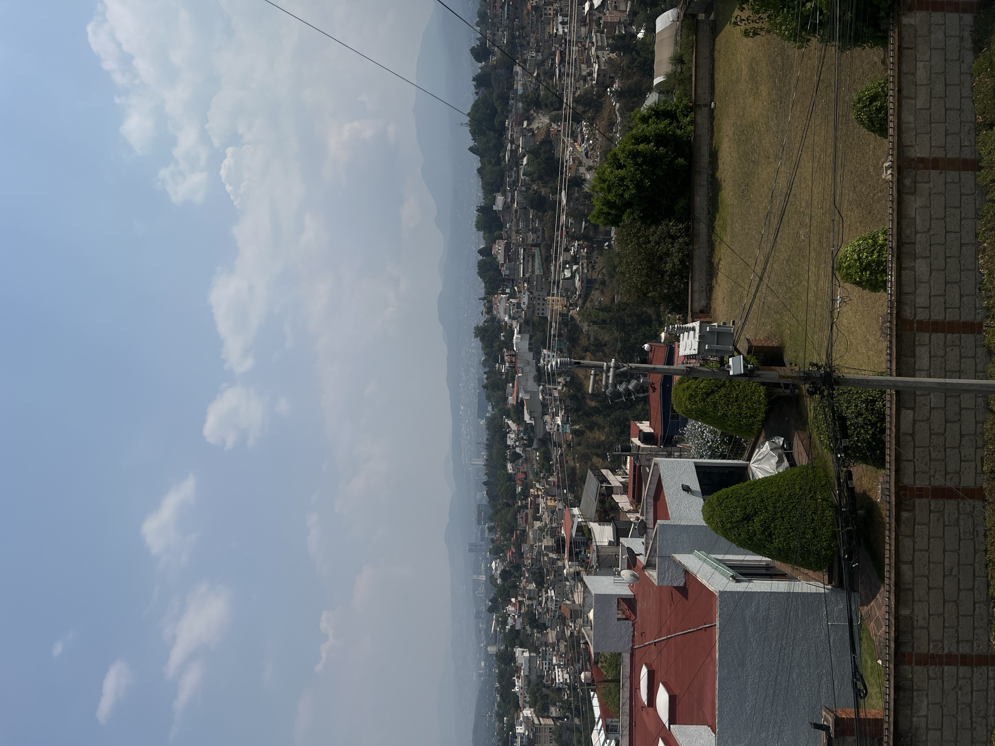 The image shows a cityscape in Mexico City, Mexico, with an expansive view of densely packed houses extending into the distance toward hazy mountains on the horizon. The sky is light blue with scattered white clouds. In the foreground are rooftops of residential buildings, distinguished by red tile roofs and white walls. A brick wall lines the edge of a property, and a grassy yard with landscaping, including trimmed bushes, is also visible. Electrical lines stretch across the scene.