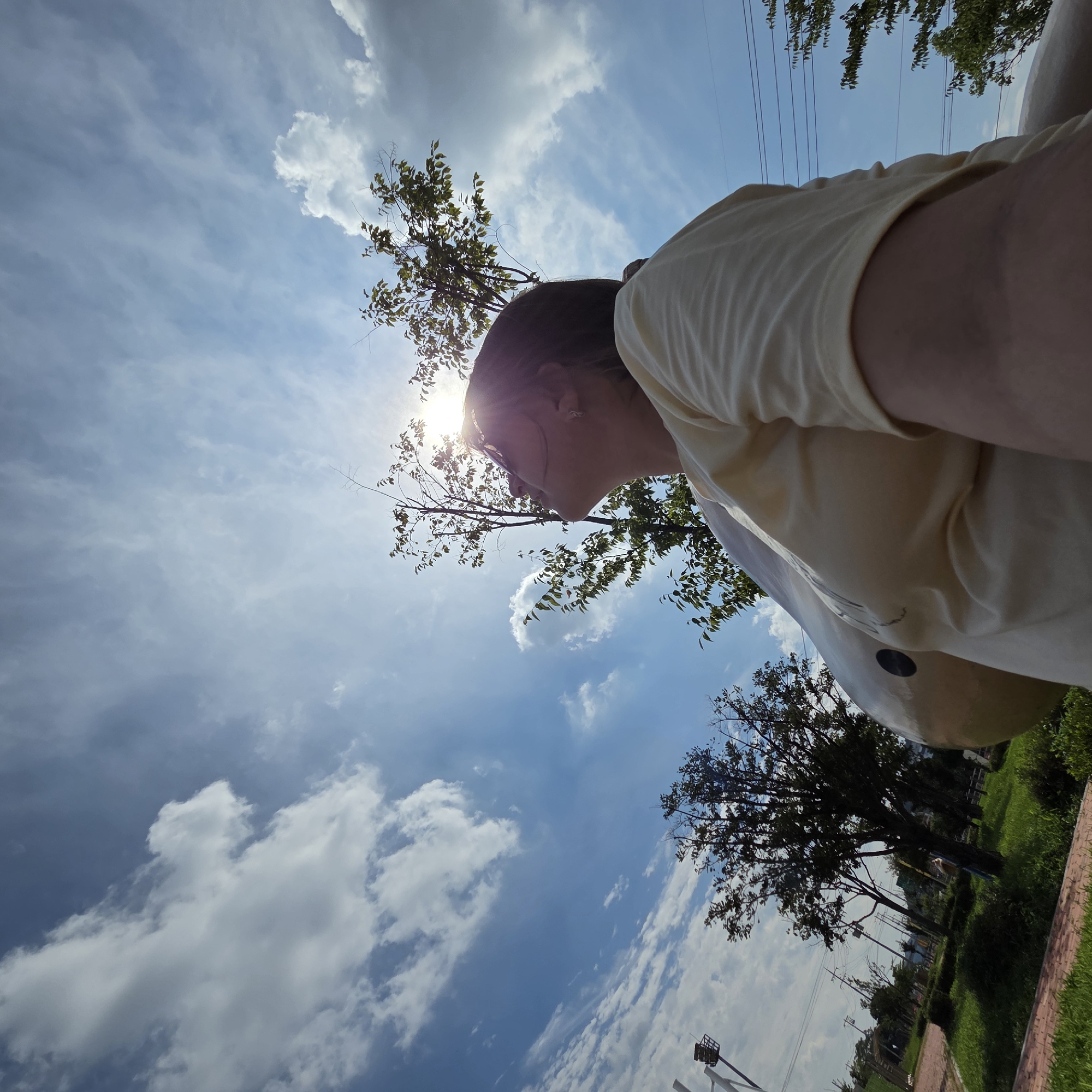 In Dunnae-myeon, South Korea, a low-angle shot captures a person wearing a light yellow t-shirt, looking up at a bright sun. The sun's rays are piercing through the leaves of a tall tree, creating a lens flare effect. The sky is a vibrant blue with scattered white clouds. The person appears to be in a park or outdoor recreational area, with lush green grass and trees visible in the background. A large, spherical sculpture is partially visible in the foreground, and power lines can be seen against the sky.