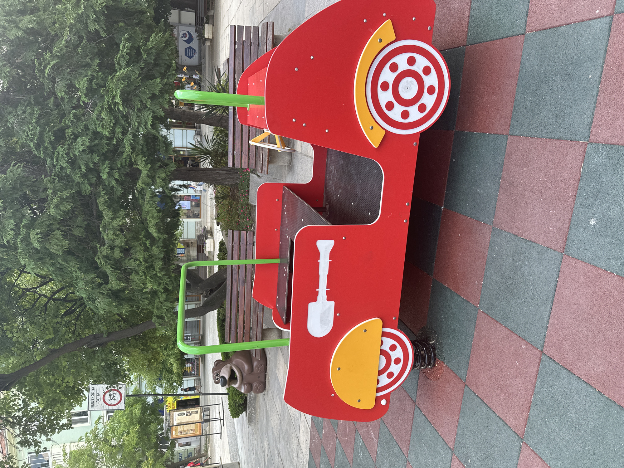 A red playground ride shaped like a car is the primary subject of this medium shot. The car has white and yellow accents on its wheels and a white shovel graphic on its side. It is situated on a patterned ground made of red and grey rubber tiles.
In the background, a large green tree with dense foliage dominates the left side of the frame. Behind the tree, a wooden park bench is visible. Further in the background, there are buildings and signs, suggesting an urban or public park setting. A brown bear-shaped sculpture is partially visible in front of the bench.
The scene appears to be outdoors during the daytime, with ample natural light. The weather seems to be overcast or the lighting is diffused, as there are no harsh shadows. The overall atmosphere is one of a public recreational space.
Visible text includes a red and white circular sign with an image and the text "ПЕШЕХОДНА ЗОНА" which translates to "PEDESTRIAN ZONE" in Bulgarian. Another sign is partially visible to the right of it.
There are no people actively engaged with the playground ride in this particular frame, but the presence of the playground and park benches indicates a place for public gathering and recreation.