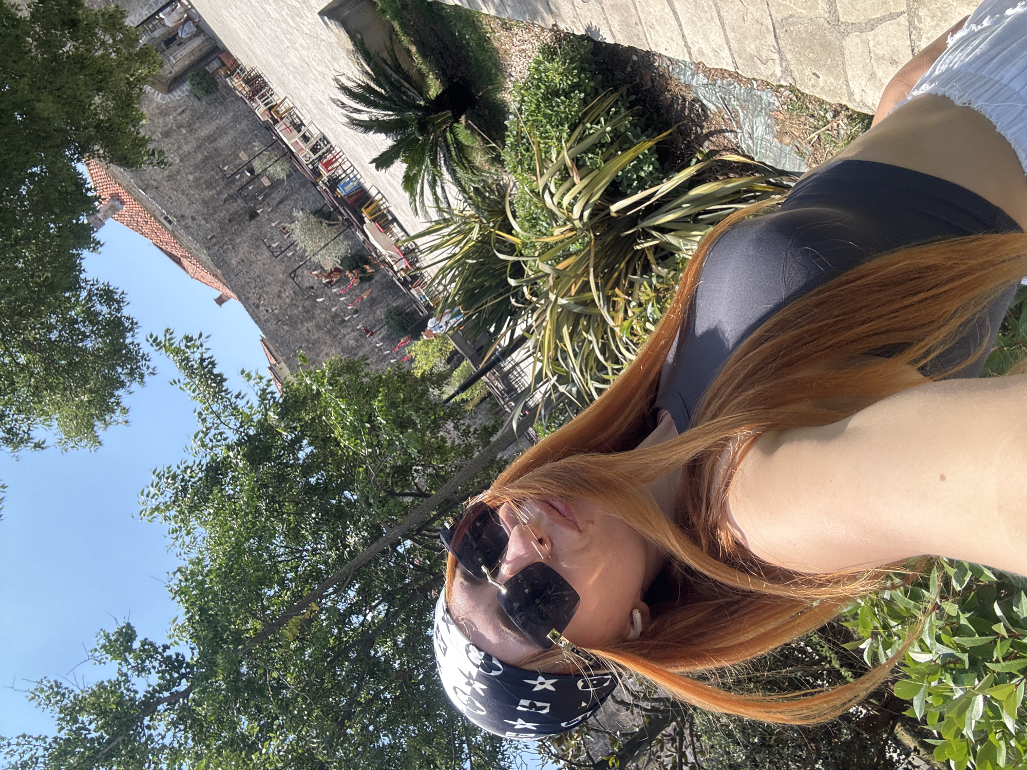 A woman with long, auburn hair, wearing a black and white bandana, large dark sunglasses, and a dark gray crop top, poses for a selfie outdoors. She has white earbuds in her ears and is looking towards the camera. In the background, there is a stone wall with a red-tiled roof, an outdoor seating area with tables and chairs, and lush green foliage. The setting appears to be a historic part of Budva, Montenegro, possibly near a restaurant or cafe within the old town walls. The sky is clear blue and the scene is bathed in bright sunlight, indicating a warm day.