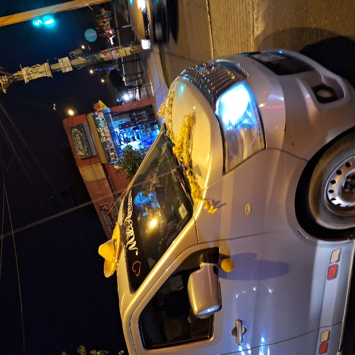 A silver car, possibly a van, sits parked at night. Its windshield is adorned with yellow tassels and a sombrero. A sticker that says "MRSAUAN" with a heart is on the door. The vehicle is in front of a building, perhaps a store, illuminated by lights. A traffic light showing green hangs above. The photo's angle is tilted.