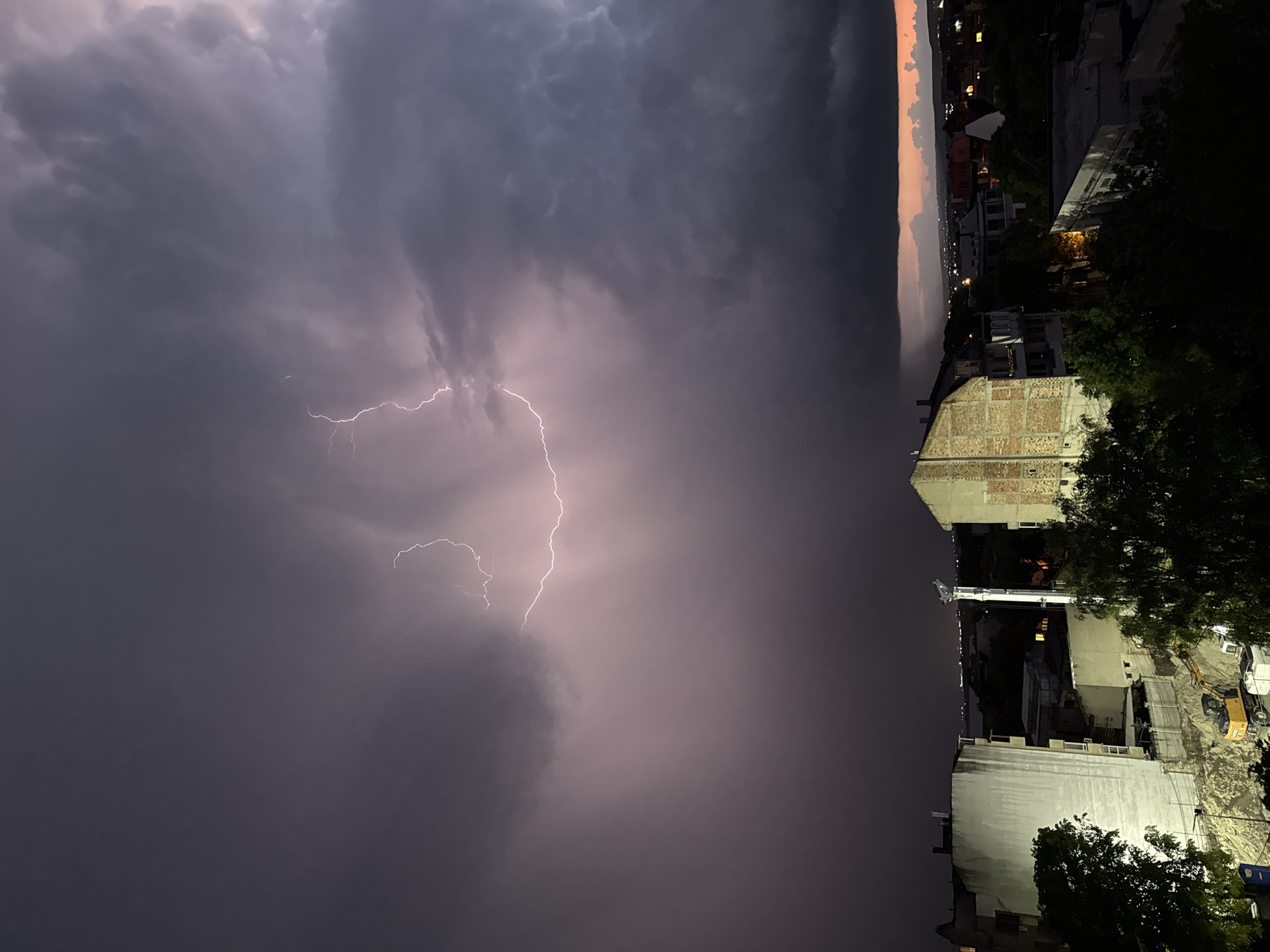 The image shows a lightning strike during a thunderstorm over Varna, Bulgaria. The sky is dark and filled with ominous storm clouds. The lightning forks down from the clouds. In the foreground, there's a view of buildings. A construction site is visible with heavy machinery. Some trees are present, adding depth to the cityscape view. The buildings appear to be a mix of residential and commercial. Further away, the horizon shows a fading sunset.