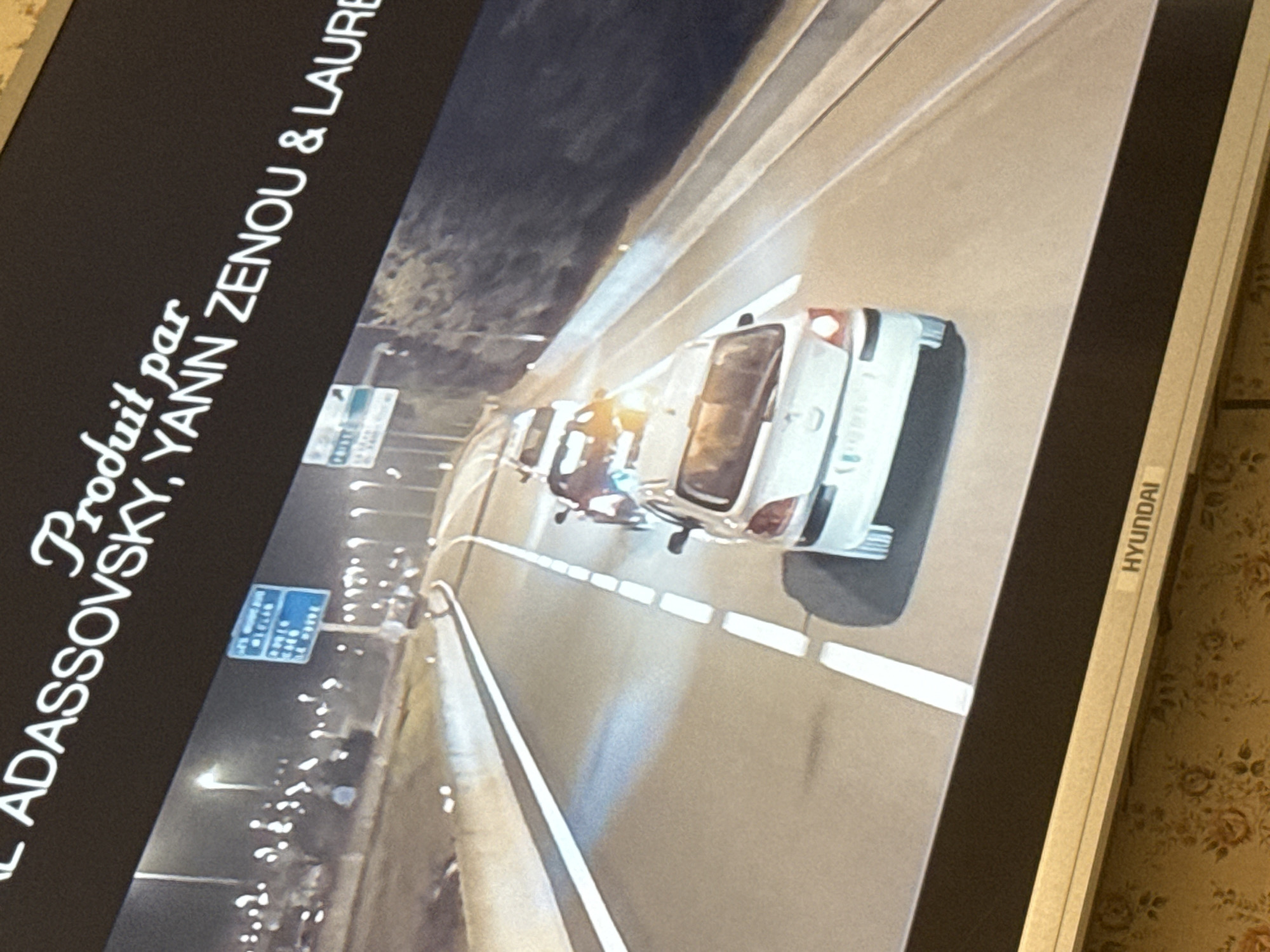 The image shows a television screen displaying a nighttime scene on a highway. Two vehicles, potentially cars or vans, are visible on the road. A figure is standing between the vehicles, possibly interacting with them. Streetlights illuminate the highway. The screen also displays text: "Produit par ... ADASSOVSKY, YANN ZENOU & LAURE". The television is a Hyundai brand. The exact location is not discernable from this image. The city of Mytishchi, Russia cannot be confirmed as the location based on the available visual information.