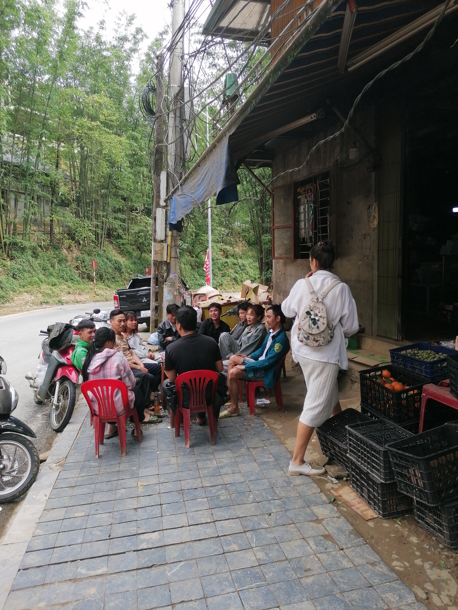 A group of people are sitting on red plastic chairs outside a building in what appears to be Sa Pa Ward, Vietnam. The group consists of approximately ten individuals, casually dressed and engaged in conversation. Some are seated, others are standing nearby. Next to the group, several parked motorbikes line the side of the road. A woman with a backpack walks towards the building's entrance. The building itself is a simple, single-story structure with a corrugated metal roof extending over the sidewalk. A tangle of electrical wires runs along the building and a utility pole nearby. A pick-up truck is parked on the road in the background. Stacks of black plastic crates, some containing fruit, are near the building's entrance. The area is paved with square tiles. Dense vegetation, including bamboo trees, is visible on a hillside behind the road. The sky is overcast, suggesting a cloudy day.