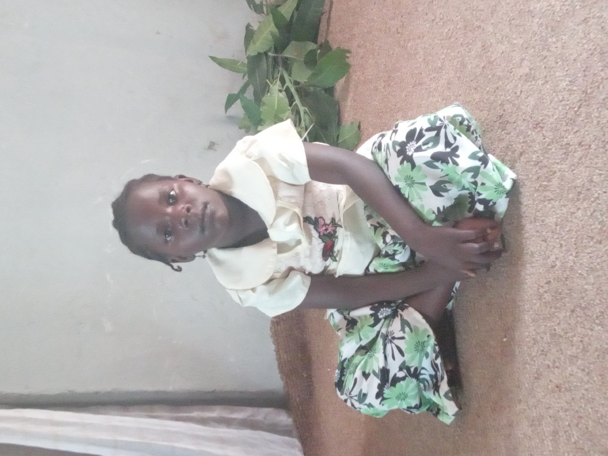 A young person with dark skin and braided hair is squatting on a tan floor. They wear a light-colored top with a collar and a floral pattern and a floral print skirt with green and white patterns. The background includes a light-colored wall and some green foliage in the upper part of the frame. The location is Sabon Tasha, Nigeria.