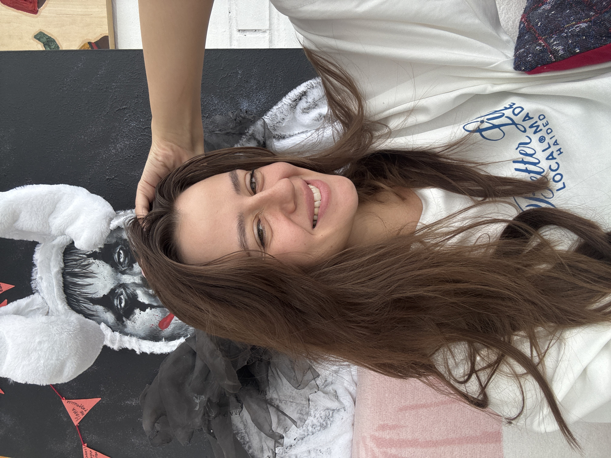 A woman is lying down on a pink and white patterned surface, with brown hair spread around her face. She is wearing a white shirt with blue lettering that reads "Locally Made." A hand is resting on her forehead. To her left, there is a black canvas with a painting of a figure wearing a white bunny hat. Below the painting are small red triangular flags strung together with string. Based on the presence of the Russian text on the flags ("Ивана не разрушила"), the location is identified as Krasnoyarsk, Russia.