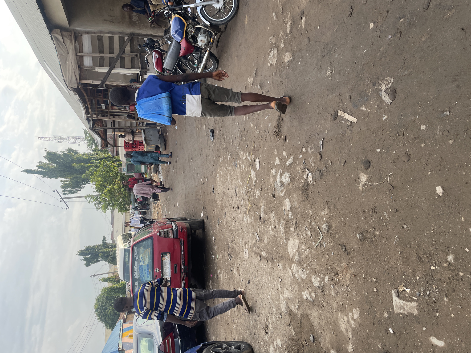 The image captures a bustling street scene, likely a market area, in Abuja, Nigeria, under an overcast daytime sky. The perspective is from slightly above, looking down a dirt road filled with activity.
In the foreground, a young man walks away from the viewer, wearing a blue t-shirt with a white stripe and dark grey shorts, carrying a light blue bag on his back. Further ahead and to the bottom right, another man, dressed in a yellow and blue striped t-shirt and grey trousers, also walks away.
The ground is unpaved, consisting of packed dirt with scattered debris, small stones, and patches of lighter soil. To the left, various market stalls or simple structures with corrugated metal roofs and wooden supports line the street. Several people are visible near these structures.
Vehicles are present, including two motorcycles parked near the market stalls towards the top of the frame. In the mid-ground, a red car with visible damage to its rear is parked, and a large white bus is partially visible behind it. In the distance, a tall metal antenna or communications tower stands against the cloudy sky. Trees and some distant buildings can also be seen.
The scene depicts a typical day in a busy urban environment, with people moving about, suggesting commerce or daily commutes. The overall impression is one of everyday life in a lively local setting.
Visible text includes "WRC" and "Volkswagen" on stickers on the rear of the red car.