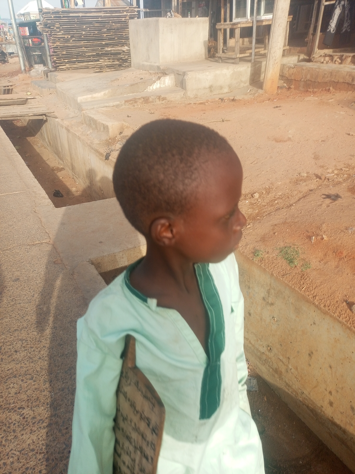 A young boy with short hair is standing outside, possibly on a street or a walkway, dressed in a light green shirt with a dark green accent on the collar. He is holding a wooden board with writing on it. He is standing in a sunny outdoor location. To his left is a drainage ditch with a concrete side. In the background, there's construction material - a pile of wood planks and what looks like parts of buildings. The location is Gombe, Nigeria.