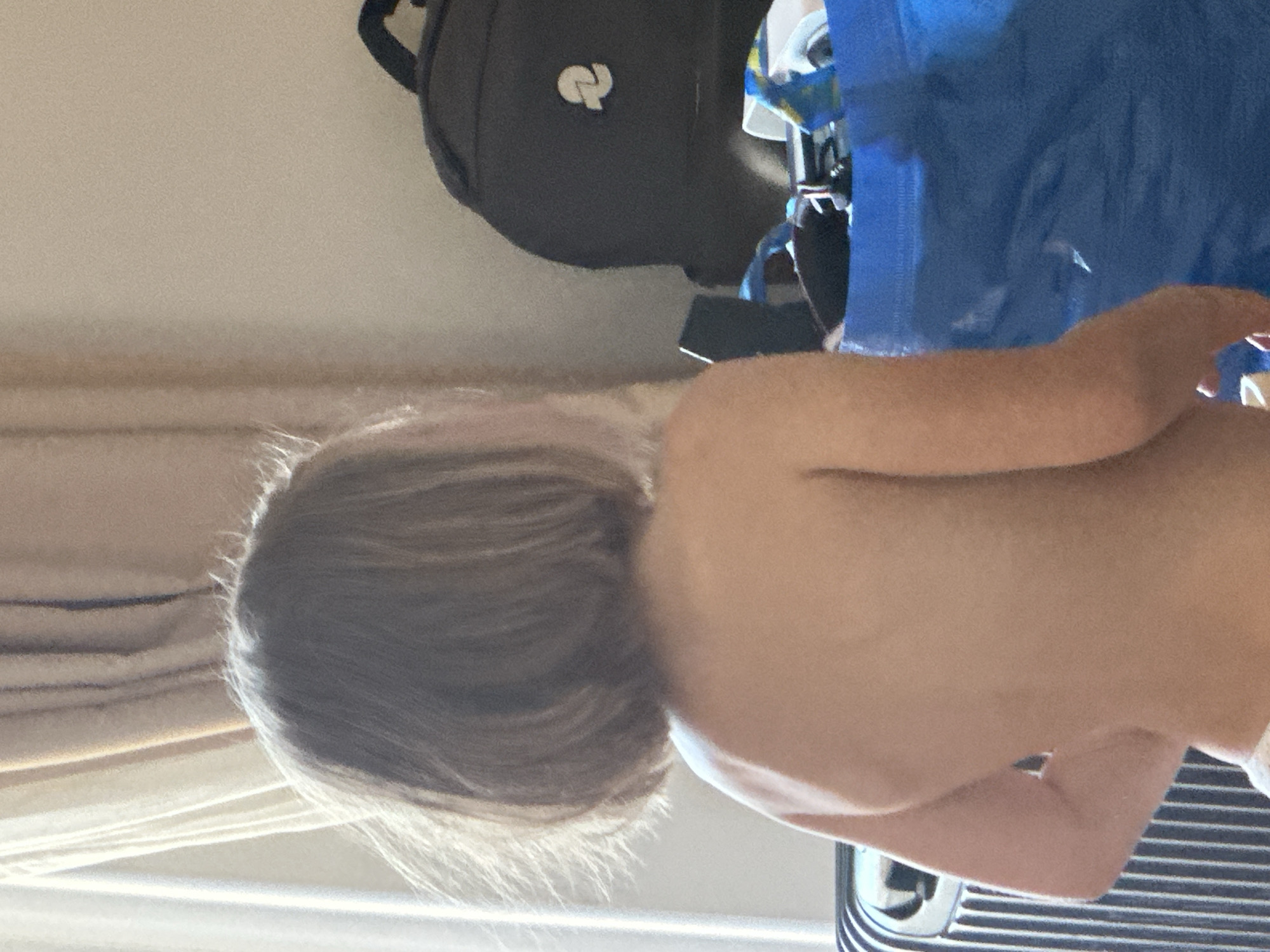 This is a vertically oriented, close-up, eye-level shot of a child from the shoulders up, seen from behind and the side. The child has light brown hair, which appears somewhat disheveled and is styled in loose waves. The child's skin is fair and appears smooth. The child is facing away from the camera and slightly to the right. The background consists of a beige wall with horizontal lines of a lighter color, possibly suggesting blinds or curtains, on the left. In the upper portion of the image, a black backpack with a white logo is visible, along with a blue object, possibly a bag or piece of luggage. The scene appears to be indoors, likely in a room. The lighting is soft and diffused, suggesting it could be daytime, though the specific time of day is not discernible. There are no clear indications of weather or strong emotions from the child, as their face is not visible. No text is clearly legible in the image.