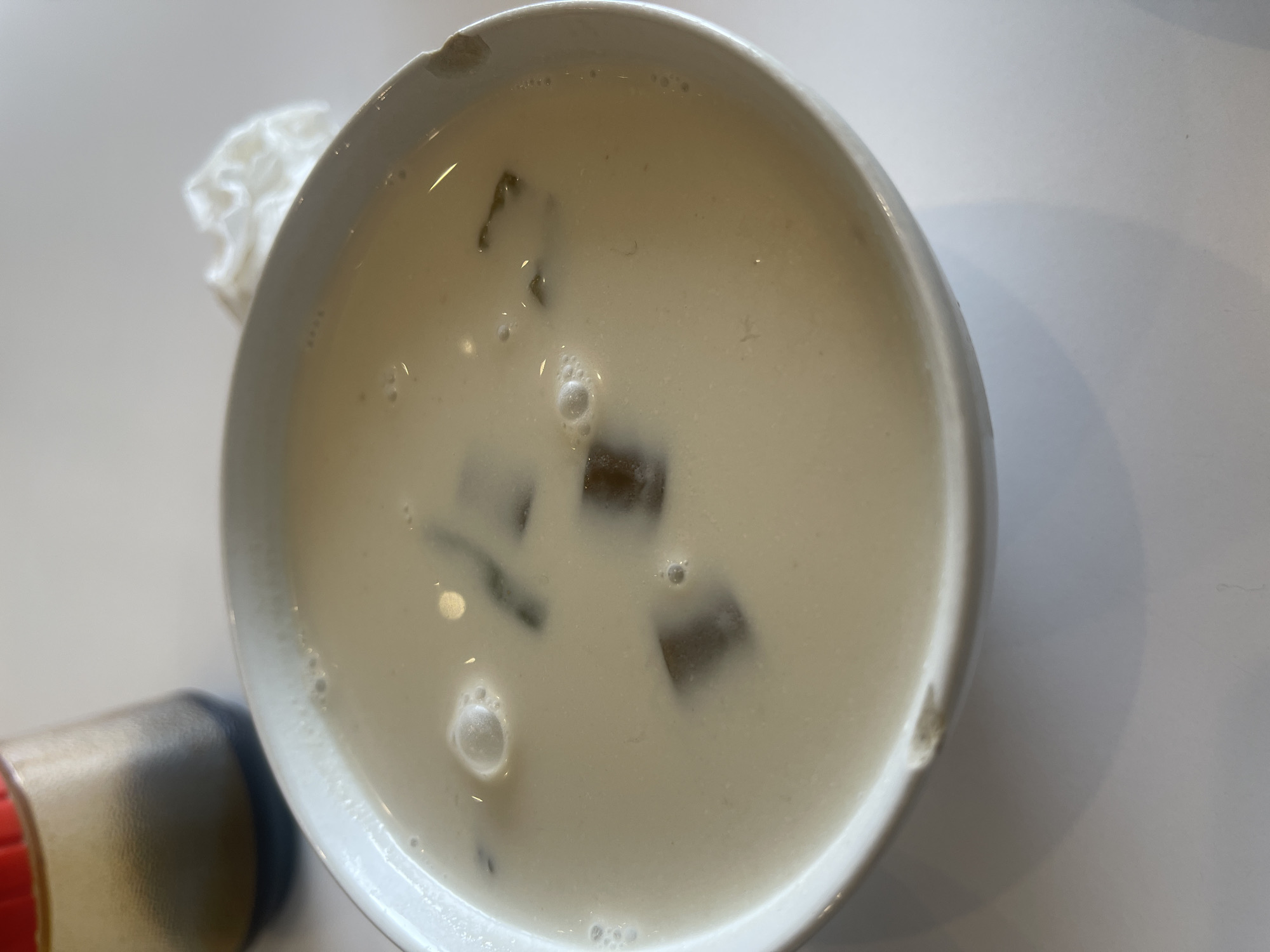 A white bowl filled with a creamy, beige liquid and dark, cubed pieces is centered in the frame, resting on a white surface. The liquid has small bubbles on its surface, and the cubes appear to be floating within it. In the upper left corner, a glass bottle with a dark liquid and a red cap is partially visible. To the upper right of the bowl, a crumpled white napkin is present. The scene is well-lit, suggesting an indoor setting. The white surface on which the bowl sits has a shadow cast from the bowl, indicating a light source from the upper right. There are no people visible in the image.