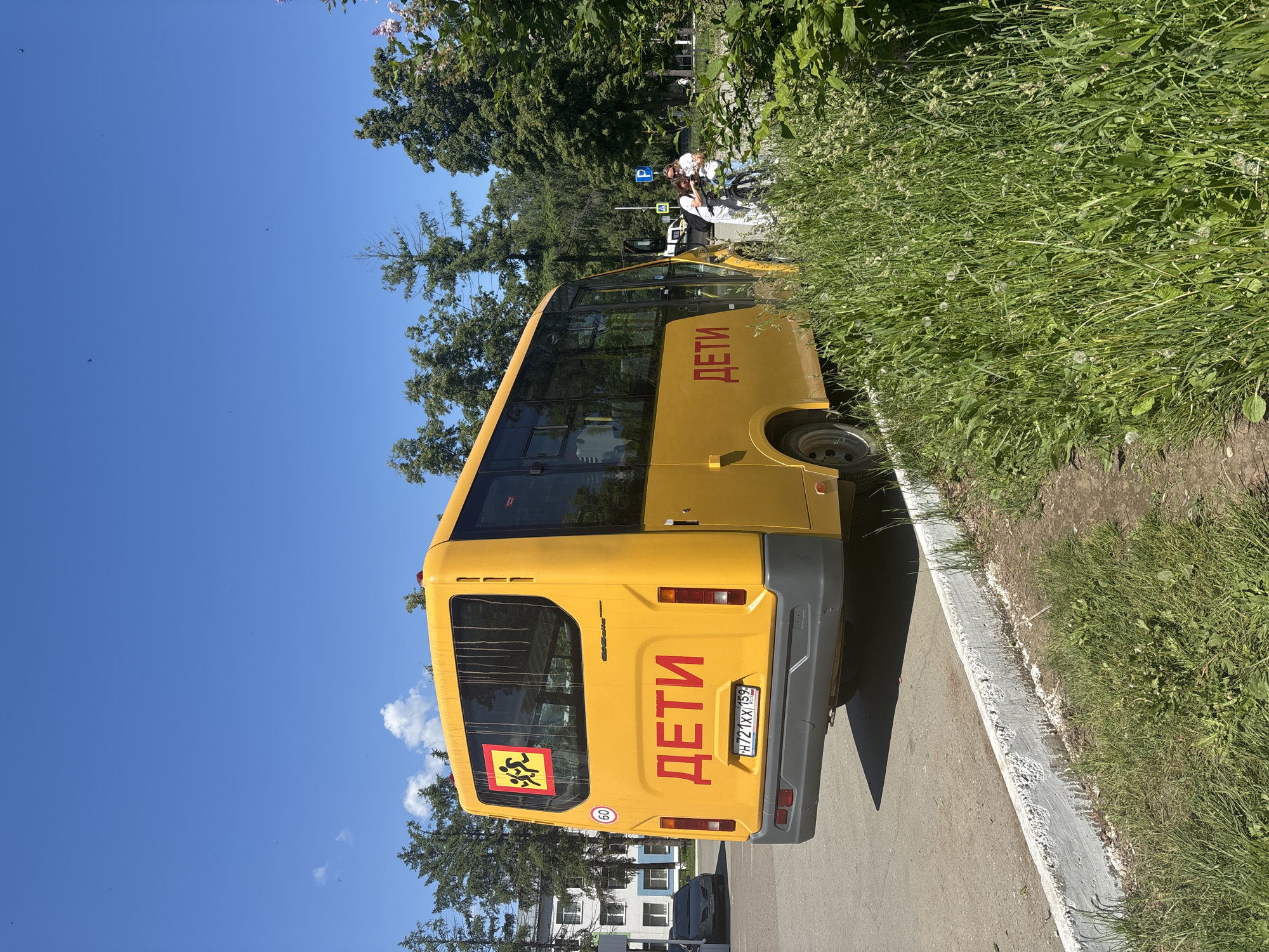 A yellow school bus, marked with "ДЕТИ" (Children) in red Cyrillic lettering on the rear, is parked on the side of a road in Chaikovsky, Russia. The bus has a speed limit sticker of "60" and a "Children crossing" symbol on its rear window. Two people, one pushing a bicycle, are visible near the bus to the right, partially obscured by overgrown grass and bushes. A dark-colored car is parked on the left side of the road. The scene is set under a clear blue sky with scattered clouds, surrounded by green trees and a white concrete curb.