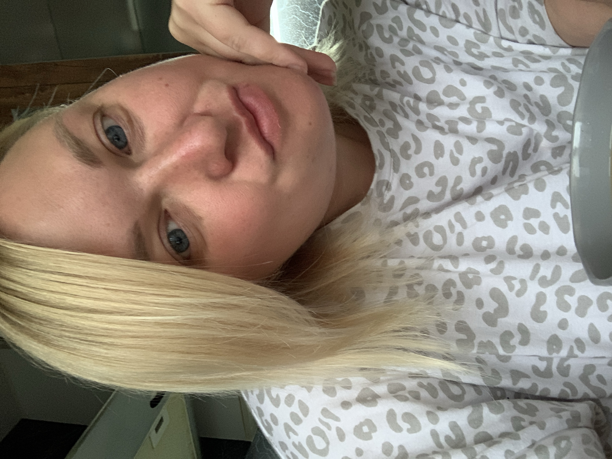 A blonde woman with blue eyes is holding her hand to her face. She is wearing a light-colored, animal print shirt. A gray object, likely a plate, is on the right. The scene is indoors. There is no information about the location being in Chaikovsky, Russia.