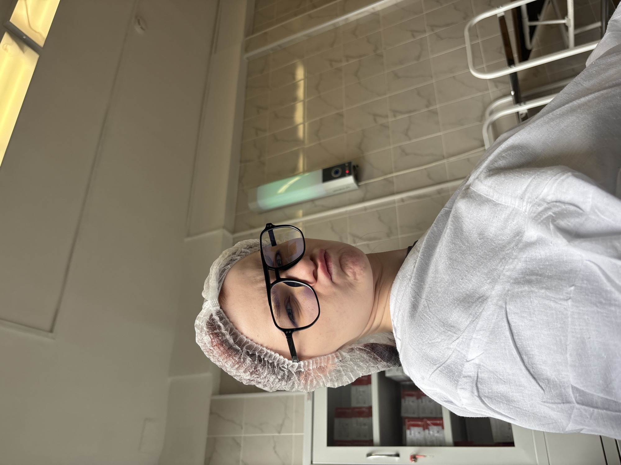 A person wearing a white disposable hairnet and a white smock is looking at the camera with a pouting expression. They are wearing large, black-rimmed glasses. In the background, there is a white cabinet filled with boxes, tiled walls, and a fluorescent light fixture on the ceiling. A vertical lamp with a green light is mounted on the wall. The scene appears to be an indoor setting, possibly a medical or laboratory environment.