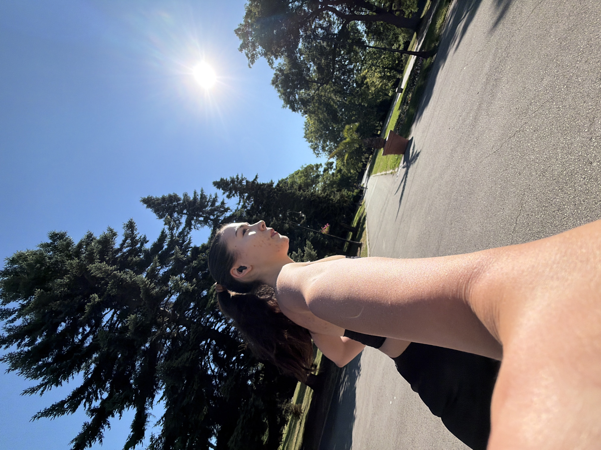 The image depicts a young woman with dark hair tied in a ponytail, wearing a black sports bra and black leggings, running on an asphalt path in a park. She has earbuds in her ears. The sun is shining brightly in a clear blue sky, casting long shadows. Mature trees with dark green foliage line the path. In the background, there is a large terra cotta pot containing a palm tree. The scene is outdoors, likely in the city of Varna, Bulgaria, given the context. The perspective suggests a first-person view, possibly from someone running alongside or filming the woman.