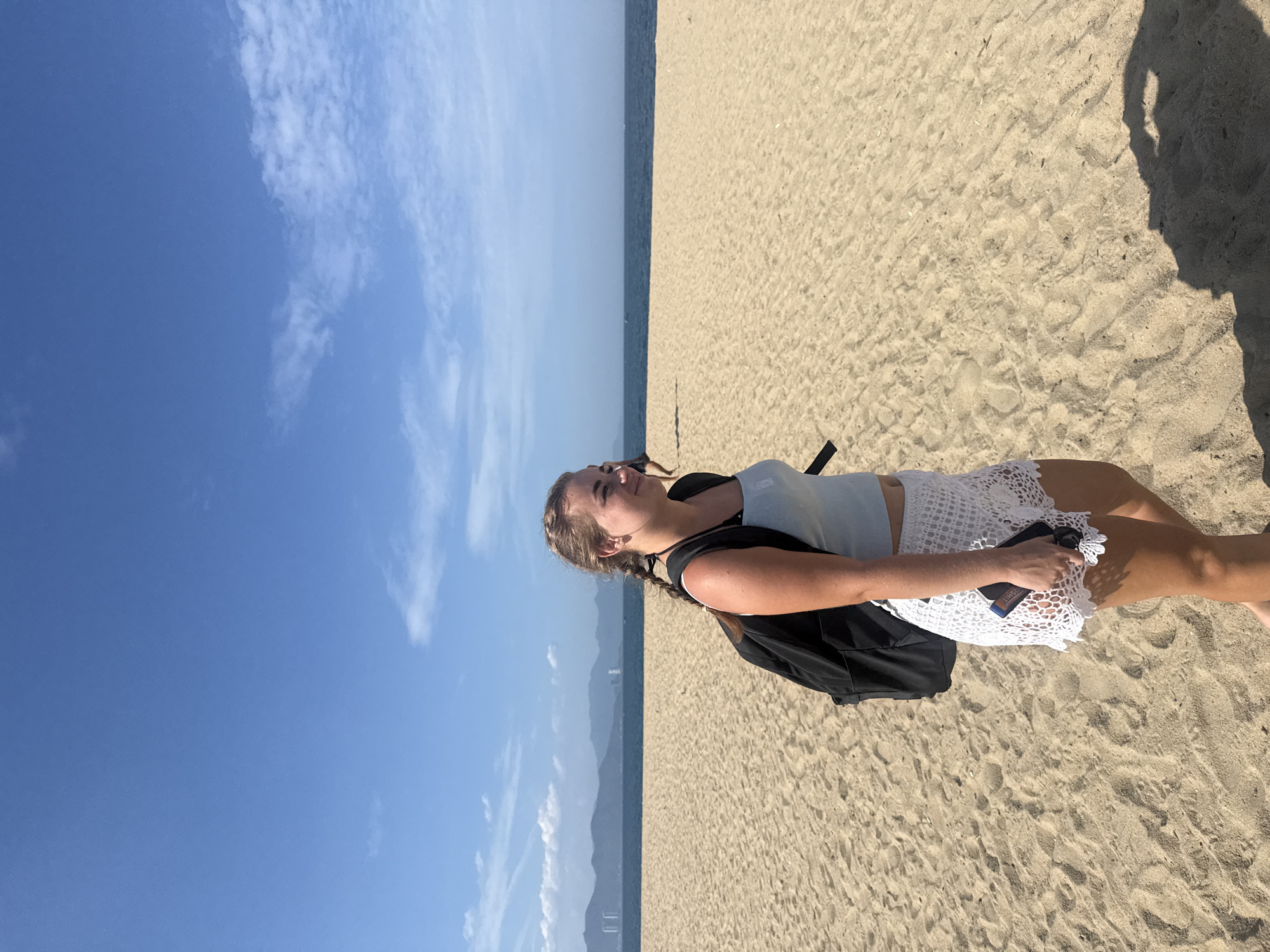 A young Caucasian woman with blonde braided hair stands on a sandy beach in Nha Trang, Vietnam. She is wearing a light blue tank top and white crocheted shorts, and a black backpack is slung over her shoulders. The woman is holding a dark-colored object, possibly a phone, in her right hand. The beach is wide and appears to be mostly empty, with gentle waves lapping at the shore in the background. Beyond the ocean, a distant coastline with mountains and buildings is visible under a clear blue sky with scattered clouds. The sun is casting a shadow of the woman on the sand to her right.