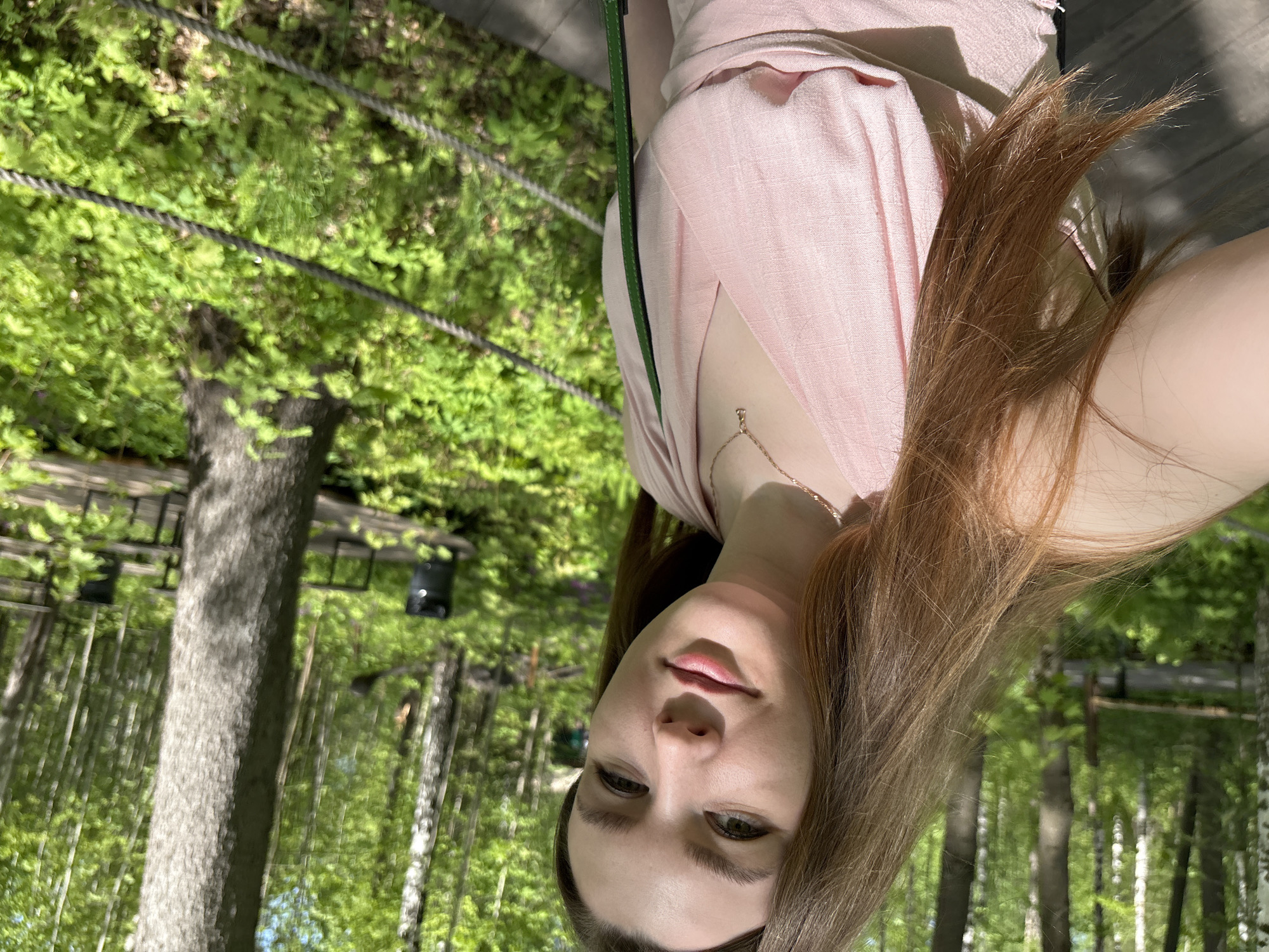 A young woman is taking a selfie. She is wearing a light pink dress. Behind her is a forest setting with birch and evergreen trees, possibly a park. There appears to be some kind of elevated platform or rope bridge structure in the background. The image may have been taken at the Gorky Park in Izhevsk, Russia.