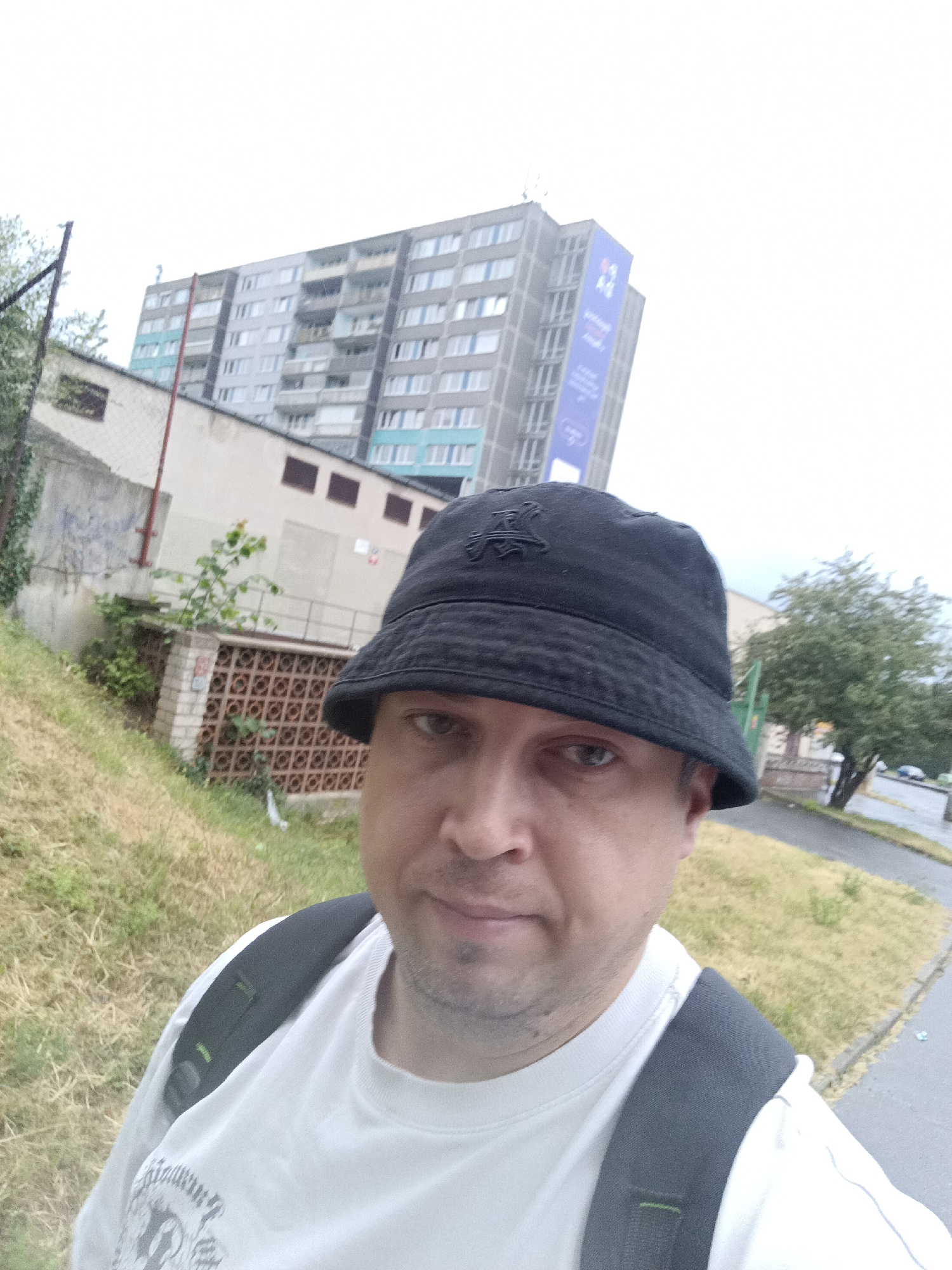 A man is taking a selfie outdoors in front of a building and a street. He is wearing a black bucket hat and a white t-shirt, with a backpack visible over his shoulders. The background shows a multi-story apartment building with many windows, a smaller building with a tiled facade, and a paved street with some parked cars. There is grass and some trees in the foreground and middle ground. The sky is overcast. The style of the apartment building, with its balconies and facade, is characteristic of socialist-era architecture common in Prague and other Eastern European cities. While the specific location within Prague cannot be determined from the image alone, the architectural style suggests it could be in one of the city's large residential districts, such as Jižní Město or Barrandov.