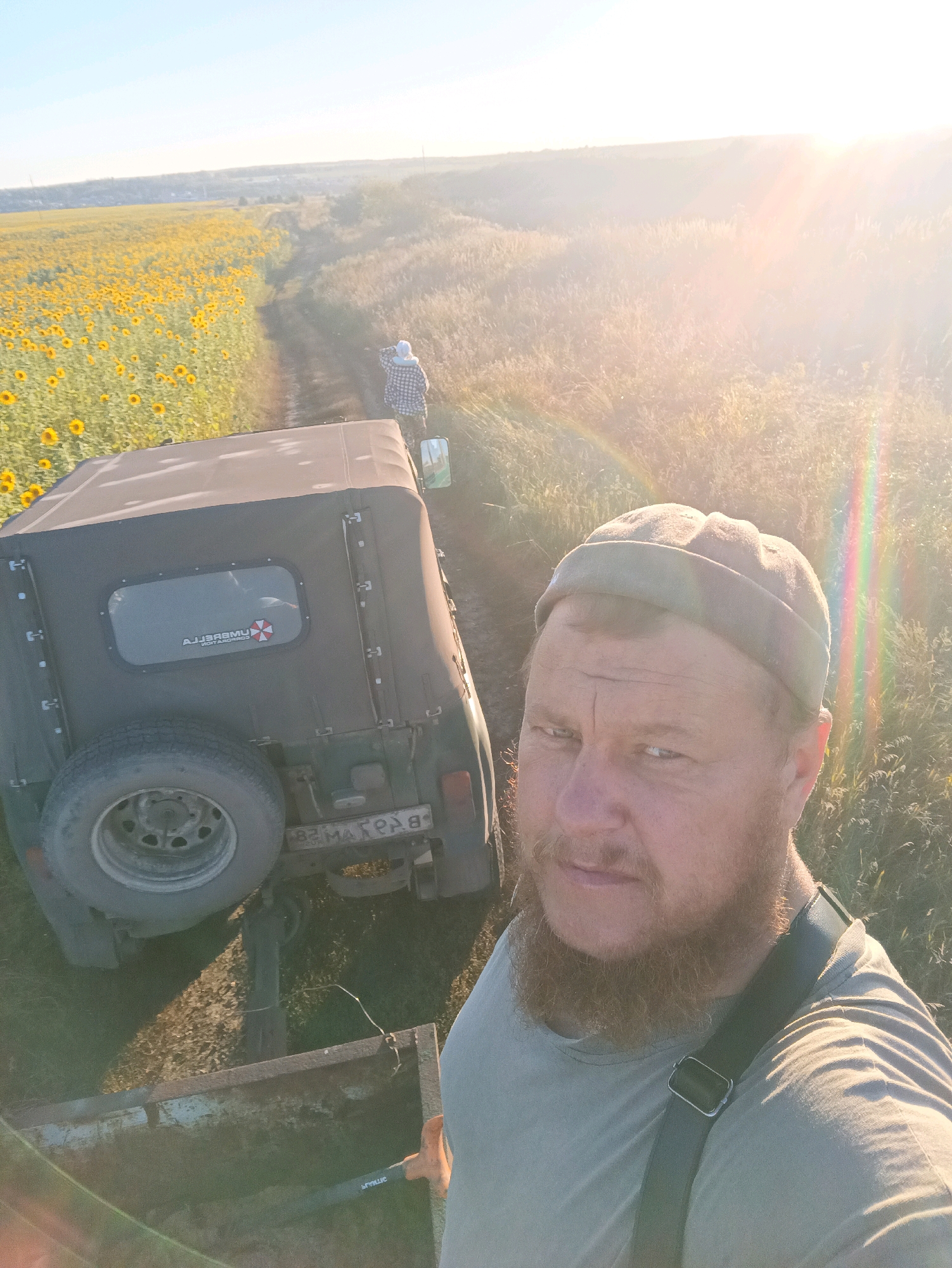 The image captures a selfie taken by a man with a beard, wearing a light green t-shirt and a dark cap, with a strap from a bag crossing his chest. He is standing in front of an olive-green off-road vehicle with a canvas top and a spare tire mounted on the back. A small trailer is attached to the vehicle.
To the left of the vehicle, a field of sunflowers stretches into the distance. A person in a checkered shirt and white hood is visible standing on the dirt road in front of the vehicle, looking towards the sunflower field. The sun is setting, casting a warm glow and lens flare across the scene, illuminating a grassy field and a distant, hazy horizon. The license plate on the back of the vehicle appears to be in Cyrillic script. The overall environment suggests a rural setting, likely during a late afternoon or early evening in Annenkovo, Russia.