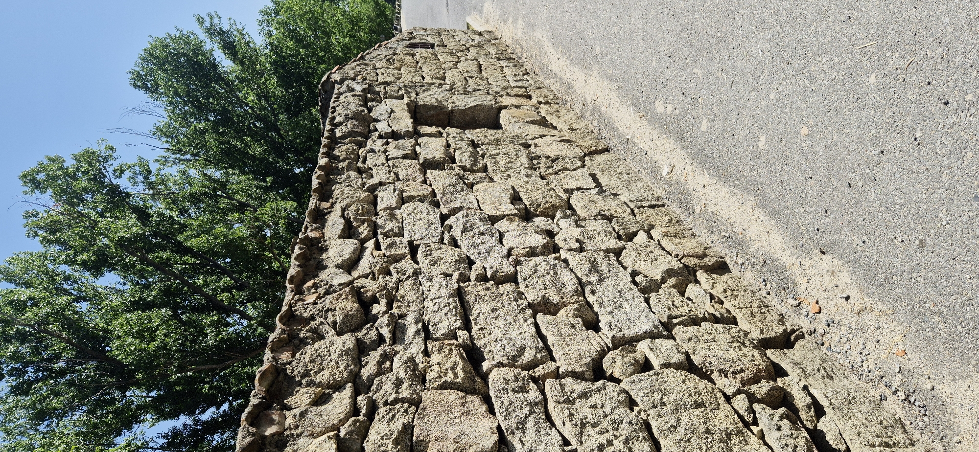 An outdoor, eye-level, vertical, medium shot captures a stone wall constructed from rough, tan-colored stones of varying sizes. The wall is dry-stacked, with visible mortar in places and is topped with smaller, reddish-brown stones. A small, dark rectangular opening is visible in the wall midway up its height. To the left of the wall is a sandy area with scattered pebbles and small pieces of broken brick. To the right of the wall is a gray asphalt road with scattered small stones and debris. In the background, a canopy of lush green trees with visible branches fills the upper portion of the image, set against a clear blue sky. The lighting suggests a sunny day with shadows cast by the wall and trees.