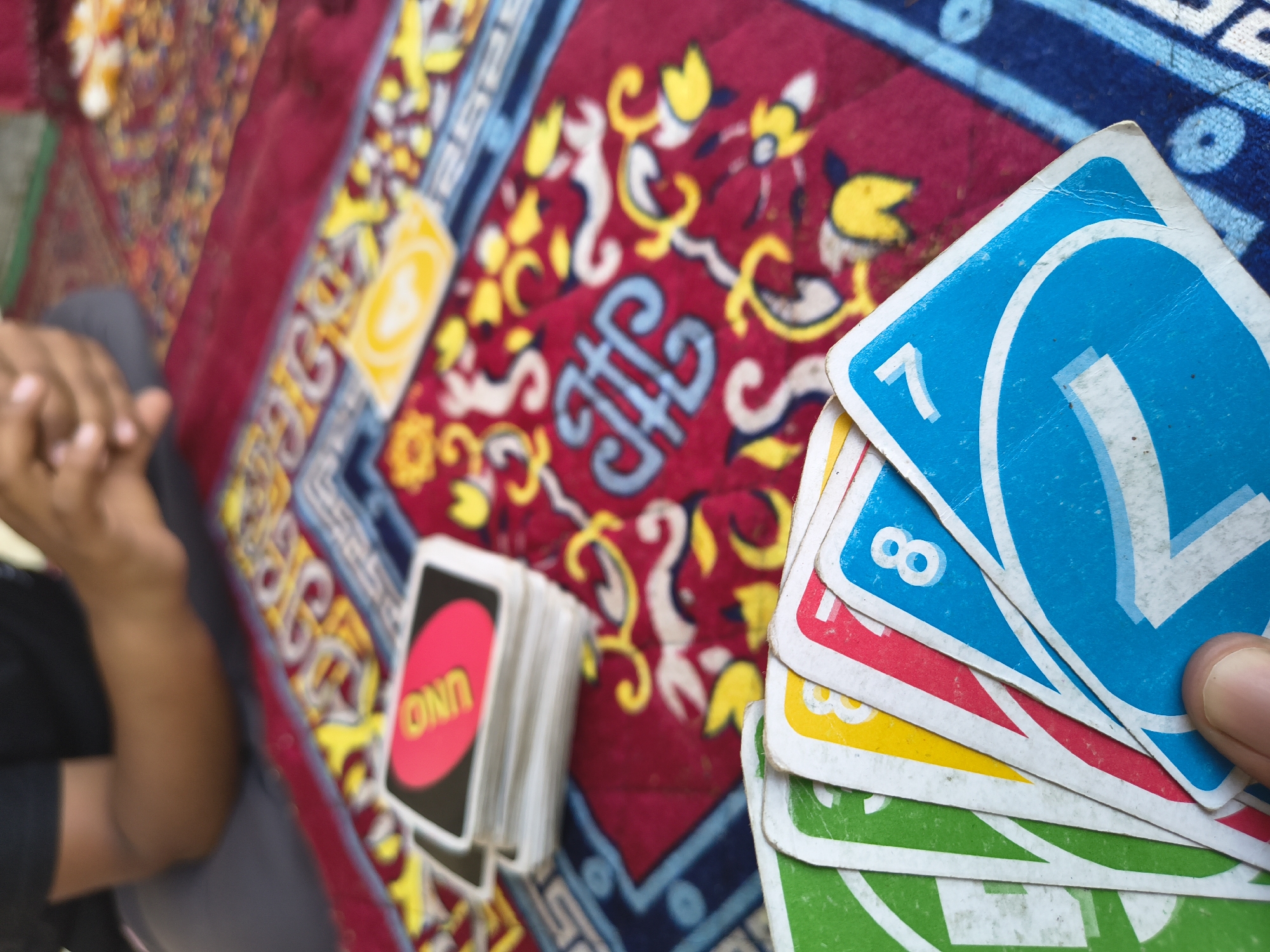The image shows a close-up of a hand holding several Uno cards, with the top card being a blue '7'. Beneath it are cards with '8' visible, and glimpses of red, yellow, and green cards. To the left is a stack of Uno cards with the 'UNO' logo visible on the top card. A person's hands and part of their torso are visible to the left of the cards. The scene is set on a patterned rug or textile with red, yellow, blue, and white designs. There is no information within the image to confirm this image was taken in the city of Haini, India.