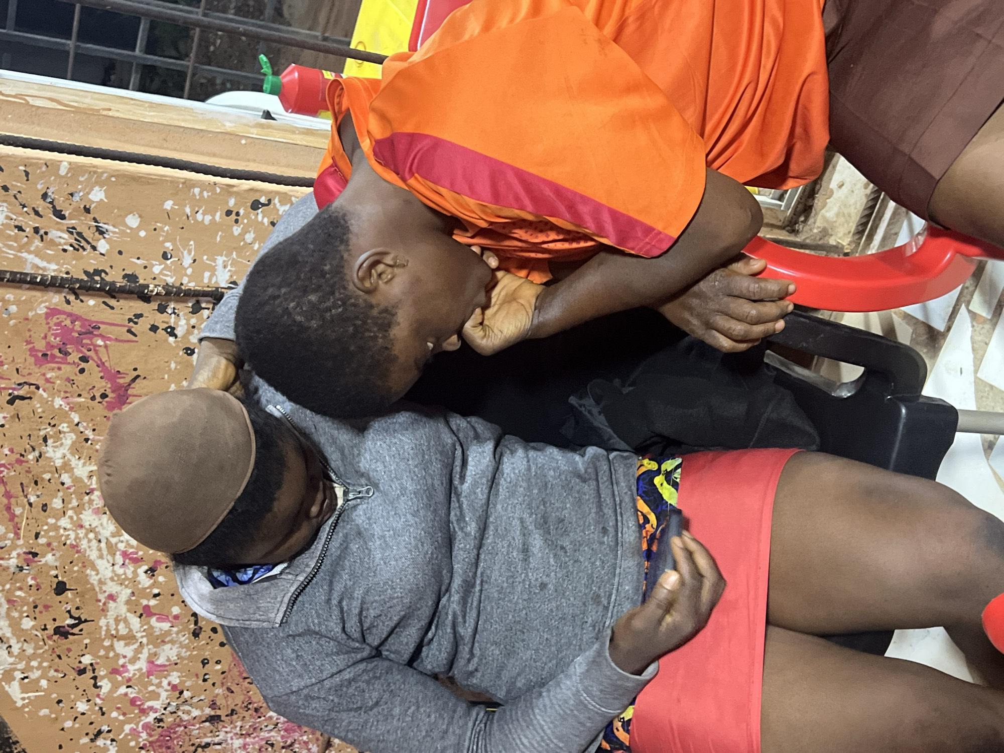 In Nkpor, Nigeria, two individuals are seated closely together. The person on the left, wearing a grey zip-up jacket over a patterned cloth and a red skirt, has a brown skullcap on their head. Their gaze is directed downwards towards their hands, one of which is holding a dark object. To the right, another individual is dressed in an orange and red short-sleeved shirt and brown shorts, with their head bowed and their hand resting near their mouth, appearing contemplative. They are both seated on chairs, one black and one red, with a textured, paint-splattered wall in the background. A red bottle with a green cap is visible to the right of the frame.