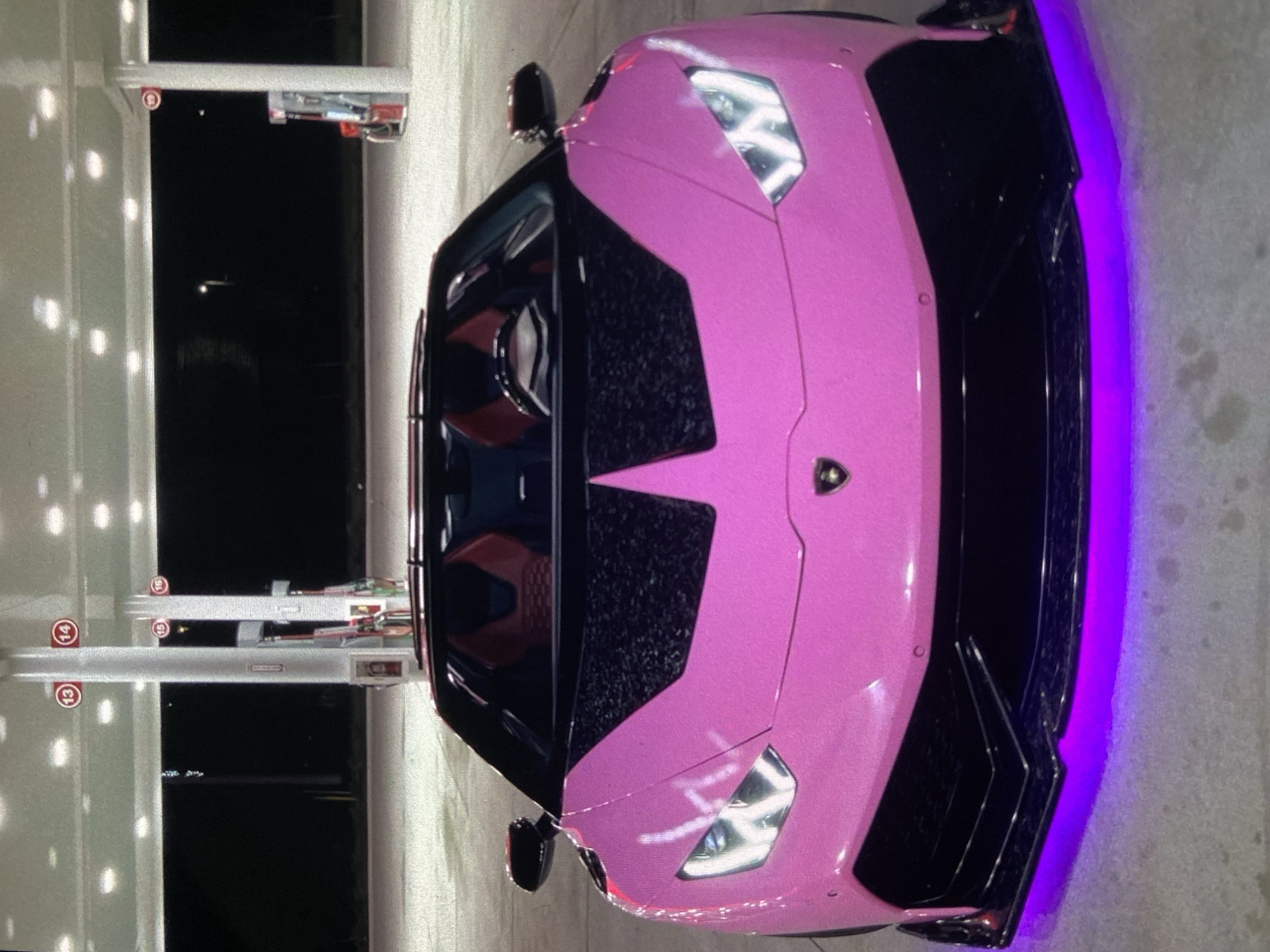 A pink Lamborghini sits parked at a gas station. Gas pump numbers 13, 14, 15, and 16 are visible. There are no people visible. The location is unknown.