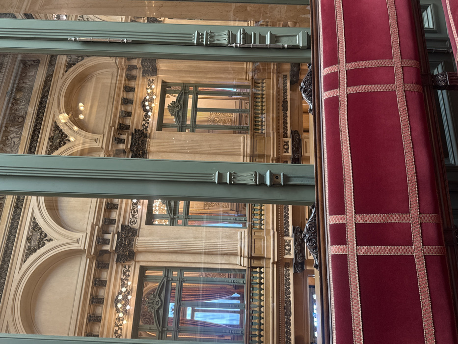The image displays a static interior scene within the Galerías Pacífico shopping mall in Buenos Aires, Argentina, captured from behind a bench. The lower third of the frame is dominated by the back of a deep red velvet upholstered bench, detailed with light gold-pink decorative trim forming a grid pattern, and featuring dark carved wooden elements on its frame.
The midground is vertically segmented by three prominent, fluted structural columns, rendered in a dark teal or emerald green hue, separating expansive glass panels. These columns exhibit classical decorative motifs at their bases and tops.
Through and reflected in these glass panels, a multi-story, highly ornate architectural interior is visible. This interior features light beige-cream walls adorned with intricate dark molding, fluted pilasters, and columns with classical capitals supporting arched openings. Upper levels display balconies with detailed dark wood balustrades. Large windows or glass doors with dark green frames are present, some partially obscured by red drapes. The ceilings and walls are illuminated by numerous small, exposed lightbulbs arranged in decorative patterns. Friezes depicting classical figures and intricate scrollwork are visible on the upper wall sections, consistent with Beaux-Arts architecture. No individuals, subjects, or actions are present in the scene.