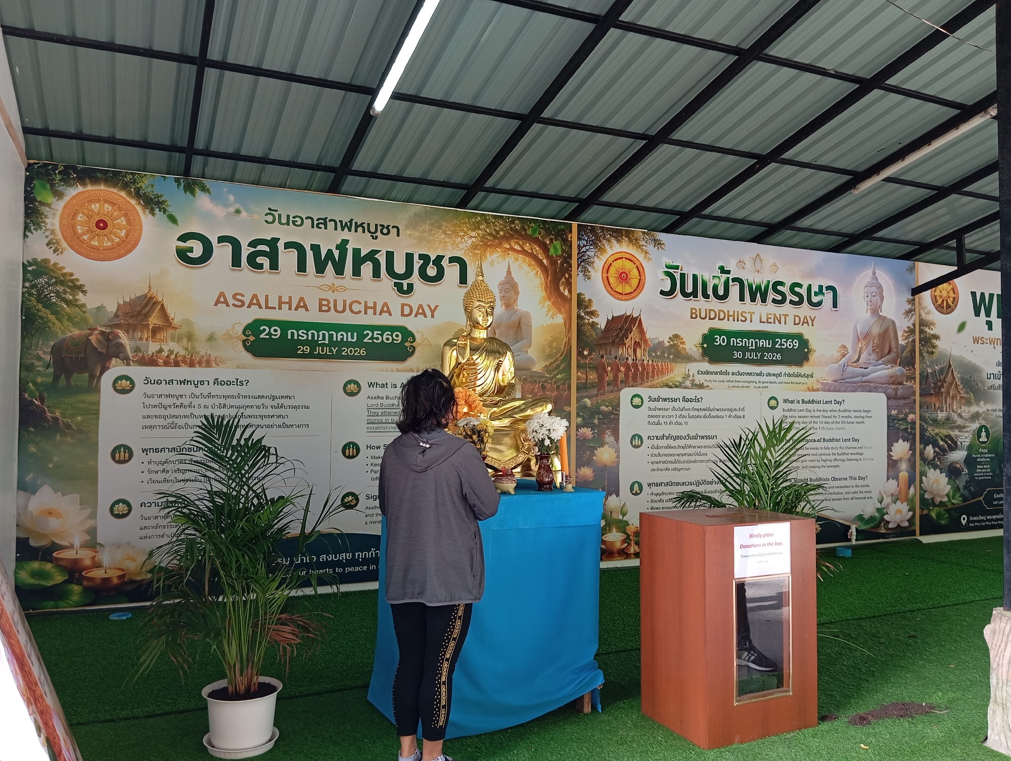 In Karon, Thailand, a woman stands facing a golden Buddha statue on an altar draped with a blue cloth. The statue is adorned with flowers and candles. Behind the altar, a large banner displays Thai and English text related to Buddhist holidays, featuring images of pagodas, elephants, and lotus flowers. The scene is set outdoors, under a corrugated metal roof supported by metal beams. To the left of the altar, a potted palm plant adds greenery to the environment. To the right, a wooden donation box with a glass window sits on the artificial grass floor. The overall atmosphere suggests a place of worship or a religious festival.