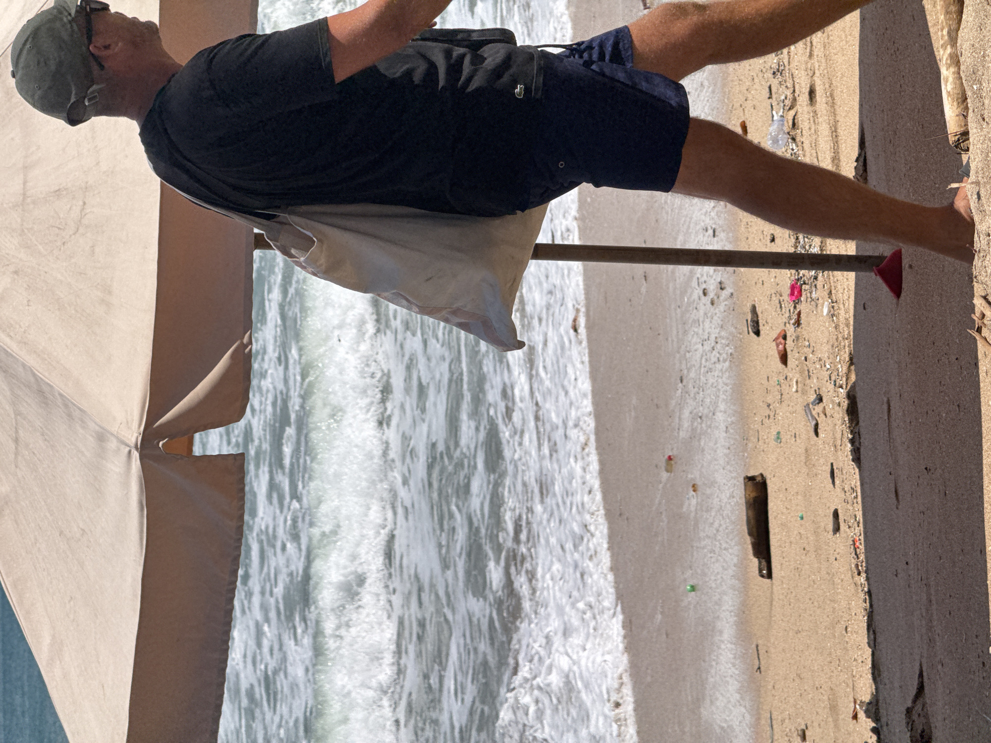 In Kuta, Indonesia, a person walks on a beach beneath a large umbrella. The person is wearing a black t-shirt, navy shorts, and a green baseball cap. They are carrying a light-colored bag over their shoulder. The beach is sandy and littered with various pieces of trash, including plastic bottles and a brown bottle. The ocean waves are visible in the background, with white foam breaking on the shore. A wooden pole supporting the umbrella is partially visible in the foreground.