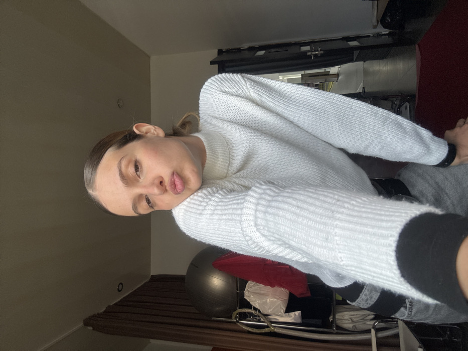 A female individual is depicted in a medium-shot selfie, positioned indoors within a room in Tbilisi, Georgia. She has light brown hair pulled back, visible freckles, and is looking directly at the camera with her head slightly tilted and lips pursed. She wears a white, ribbed knit, high-neck long-sleeve sweater and grey pants with black horizontal bands. A black wristband is visible on her left wrist.
The background on the left side shows a dark brown curtain, a dark grey exercise ball, and a metal clothing rack holding a white garment, a bright red garment, and other darker clothing items. The ceiling features a single recessed light fixture. On the right side, a dark-framed doorway is partially visible, leading to a brighter, unidentifiable space beyond. A segment of a dark red mat or rug is present on the floor in the bottom right corner. Lighting suggests an ambient, possibly natural, light source, with some shadows present.