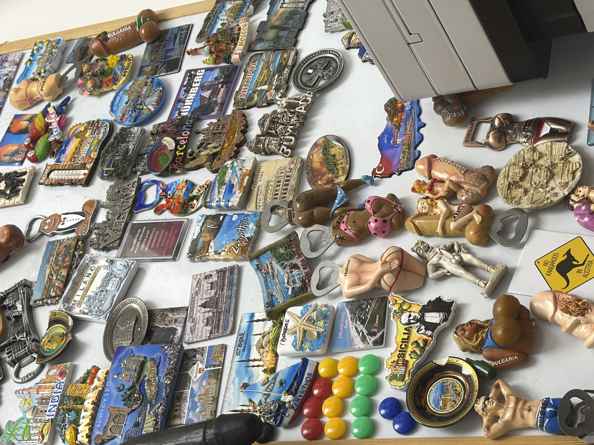 The image displays a collection of novelty magnets, primarily souvenir magnets featuring landmarks and iconic imagery from various international locations. The magnets are arranged on a white surface, likely a refrigerator or a magnetic board.
Several magnets depict famous landmarks such as the Colosseum in Rome, the Eiffel Tower in Paris, and structures from cities like Milan, Istanbul, and Budapest. There are also magnets with humorous or adult-themed designs, including figures of people and phallic shapes. Some magnets have text indicating their origin, such as "Milano," "Paris," "Nurnberg," "Hungary," "Bulgaria," "Sicilia," "Zakynthos," and "Dundee."
Specific details observed include:
- A magnet shaped like a human torso with exaggerated female anatomy, labeled "Mallorca."
- A magnet featuring a man in a tuxedo with a gingerbread man design.
- Several spherical magnets in red, yellow, green, and blue.
- A magnet with an Australian warning sign depicting a kangaroo, labeled "NO KANGAROOS IN AUSTRIA."
- A magnet shaped like a stylized female torso with the word "BUDA" and "HUNGARY."
- A magnet with a map of Sicily featuring an image of Al Pacino as the Godfather.
- A magnet with a Turkish flag and a landscape depicting the Hagia Sophia.
The location in the city of Sofia, Bulgaria, is not explicitly identifiable within the image. The scene is a close-up of a collection of decorative magnets, devoid of any context that would place it within Sofia or any other specific geographic location. The objects are static and do not depict any actions or interactions. The environment appears to be indoors, with a plain white background.