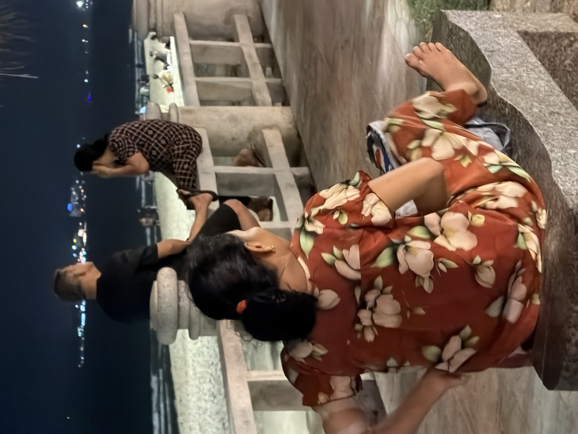 At night, four people are visible sitting and standing near a beach in Nha Trang, Vietnam. In the foreground, a woman with dark hair pulled back in a bun, wearing a red dress with a floral pattern, sits on a stone bench facing away from the camera. Her left arm is bent at the elbow with her hand on her hip. Her right leg is extended forward, with her bare foot resting on the bench. To her left, partially visible, is another person with dark hair.
Behind the woman in the foreground, a man with short gray hair is seated on a concrete ledge, looking to his right, out towards the ocean. He is wearing a black t-shirt. To his right, a woman with dark hair tied back, wearing a dark patterned dress, is seated on the same ledge, facing away from the camera. She appears to be touching her ear.
Further in the background, the ocean is visible with waves, and beyond that, a city skyline with lights can be seen. The beach sand is illuminated, and a white concrete railing runs along the edge of the promenade where the people are seated. Palm fronds are visible hanging into the frame from the top right. The overall lighting suggests it is nighttime.
