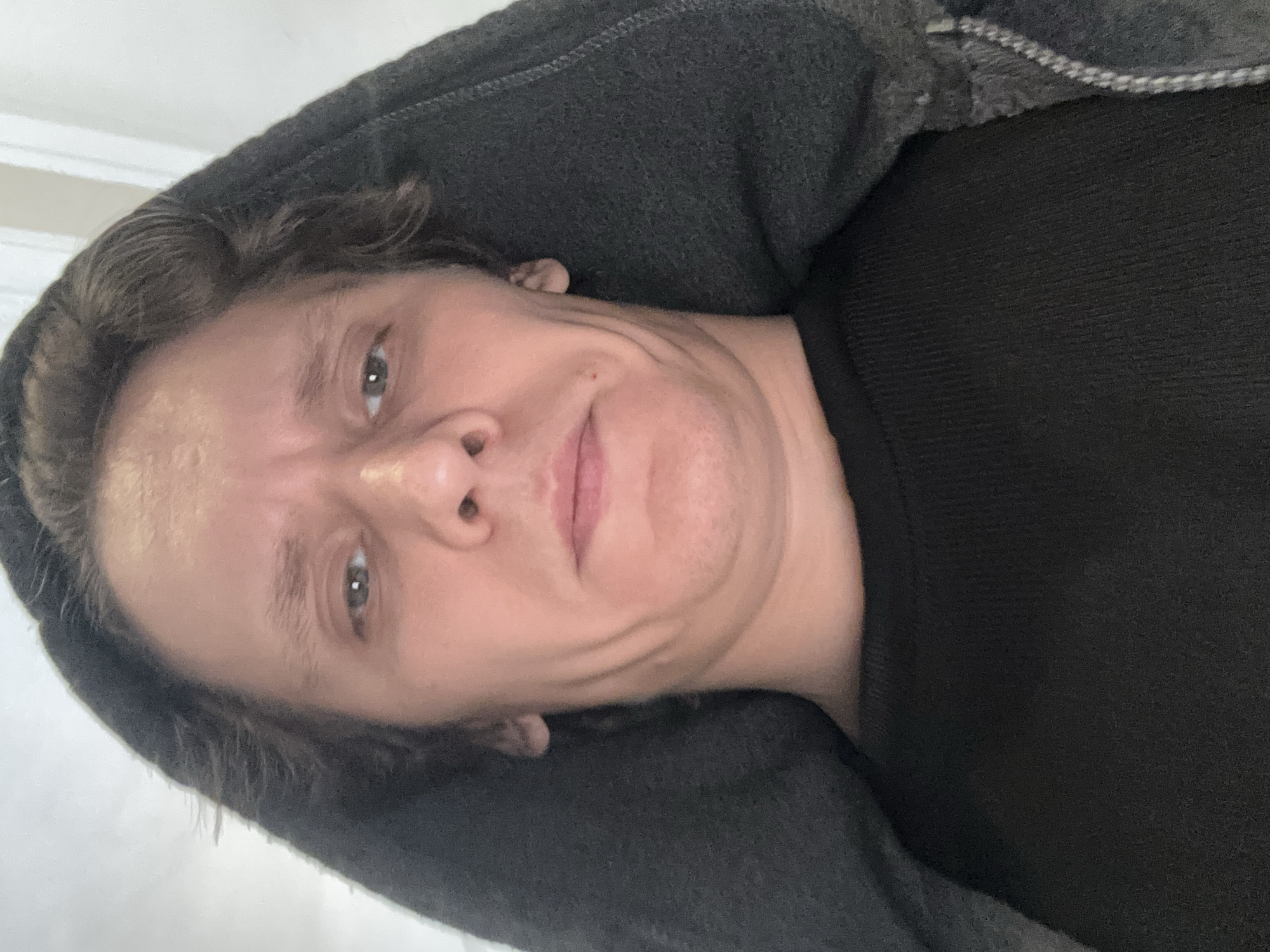 A low-angle, close-up shot shows the face of a person wearing a dark hooded sweatshirt. The person's eyes are open and looking directly at the camera. Their forehead is furrowed, and their mouth is slightly open. The hood of the sweatshirt is pulled up, framing the face. The background is blurred and appears to be an indoor setting, possibly a room with a white wall or ceiling. No specific location in San Francisco can be identified from the image.